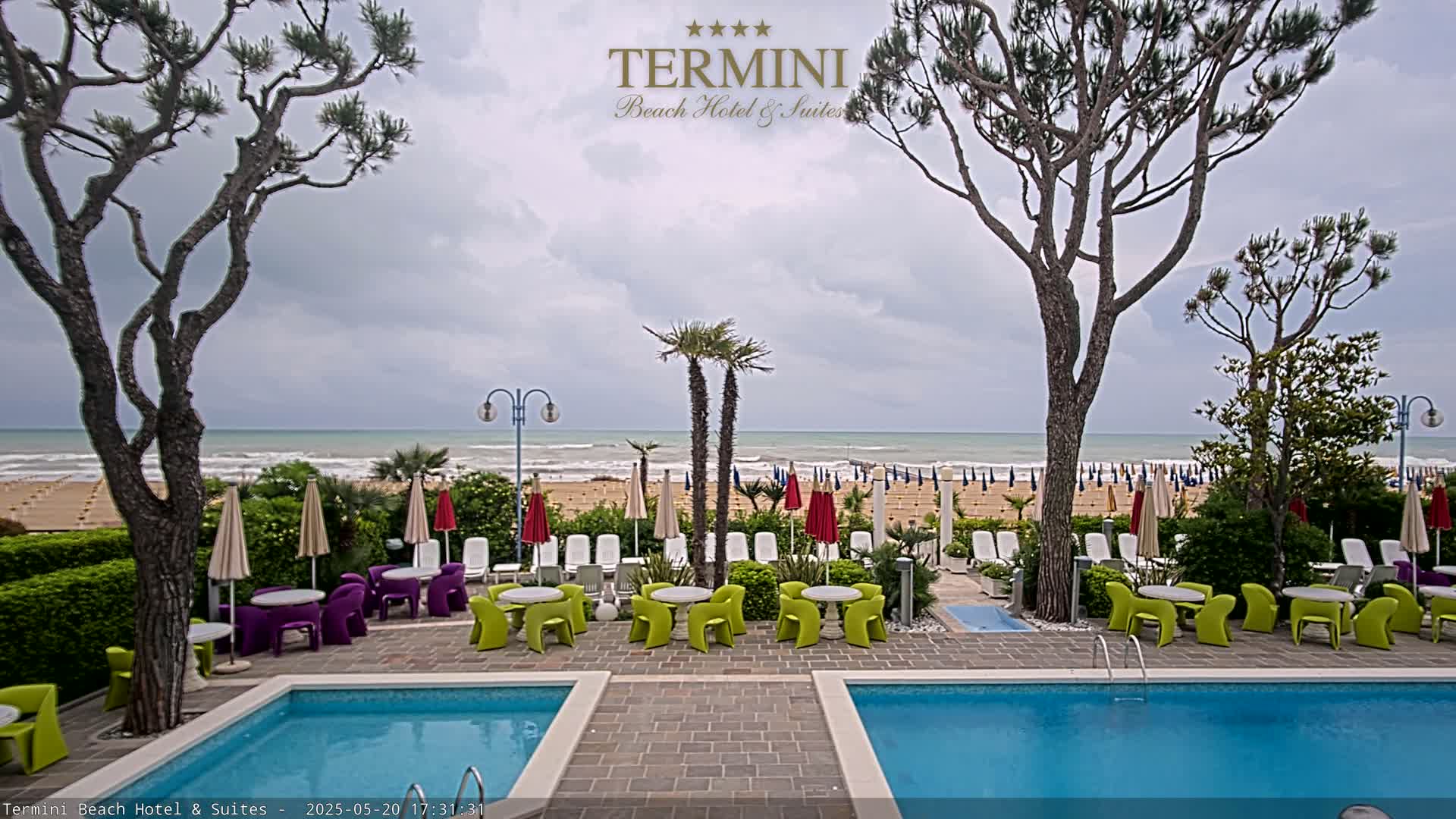 A hotel pool area with colorful furniture overlooks a beach with numerous umbrellas under an overcast sky.