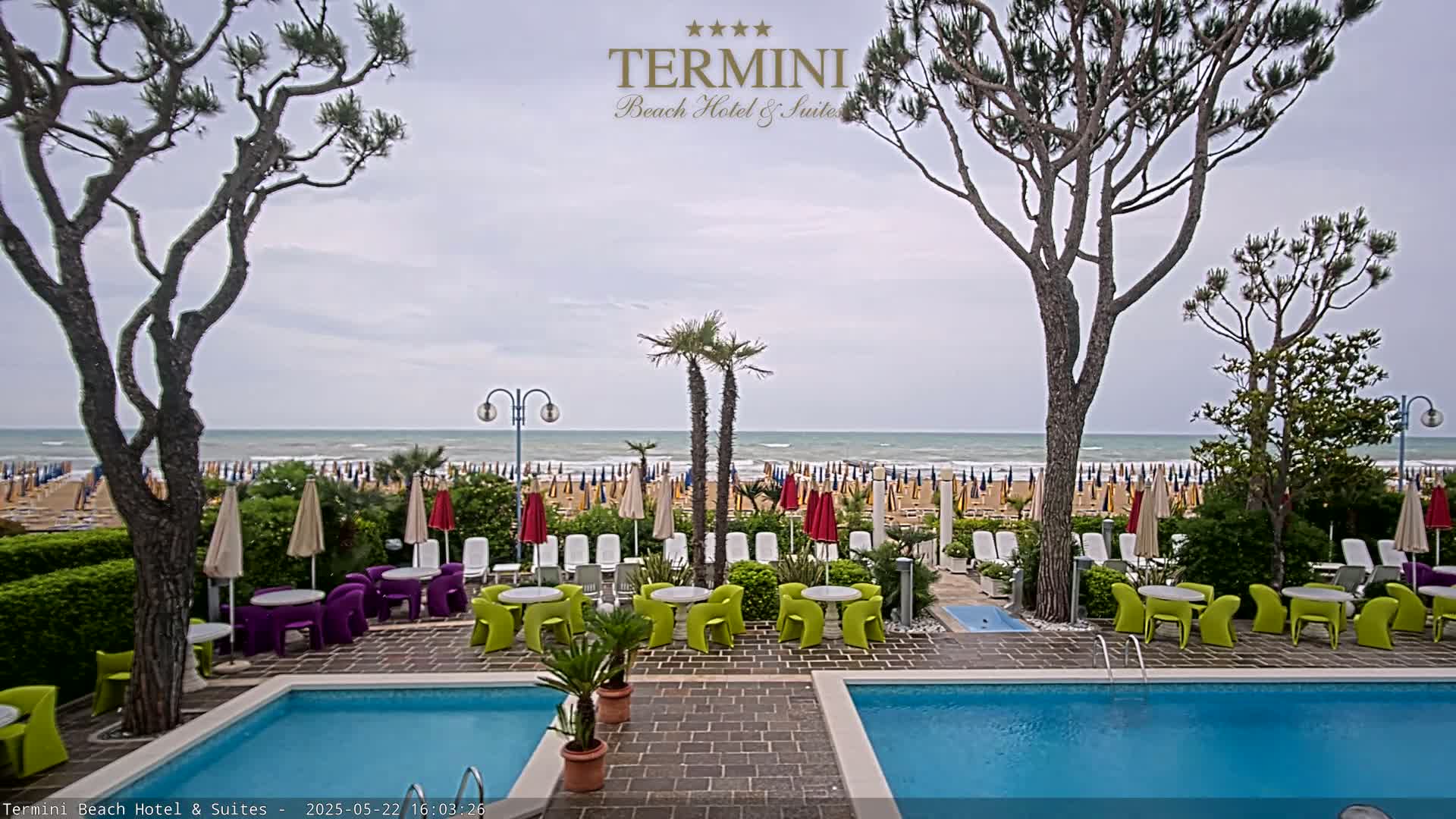 A swimming pool sits between two rows of brightly colored patio furniture, overlooking a beach with numerous umbrellas under a cloudy sky.