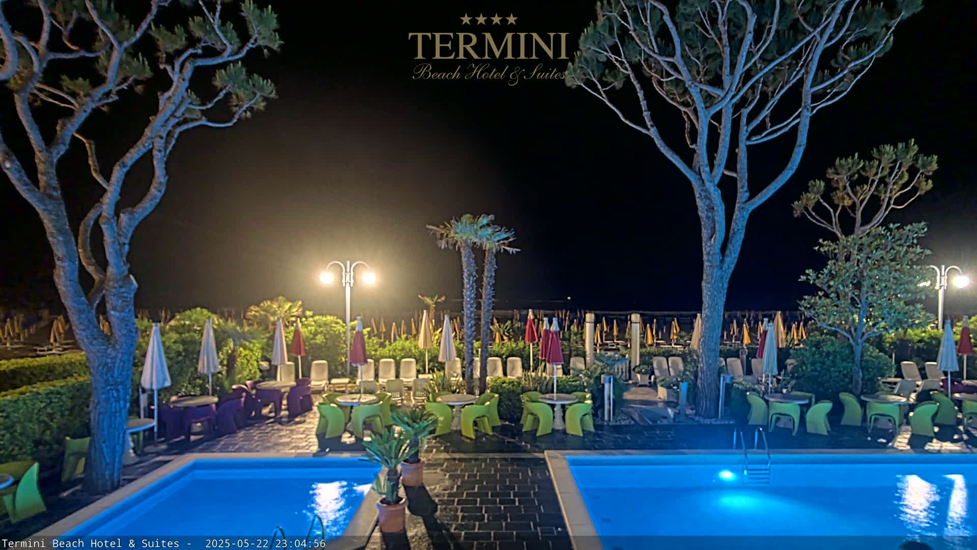 Two illuminated swimming pools sit beside a patio area with colorful chairs and umbrellas at night, under the cover of trees.