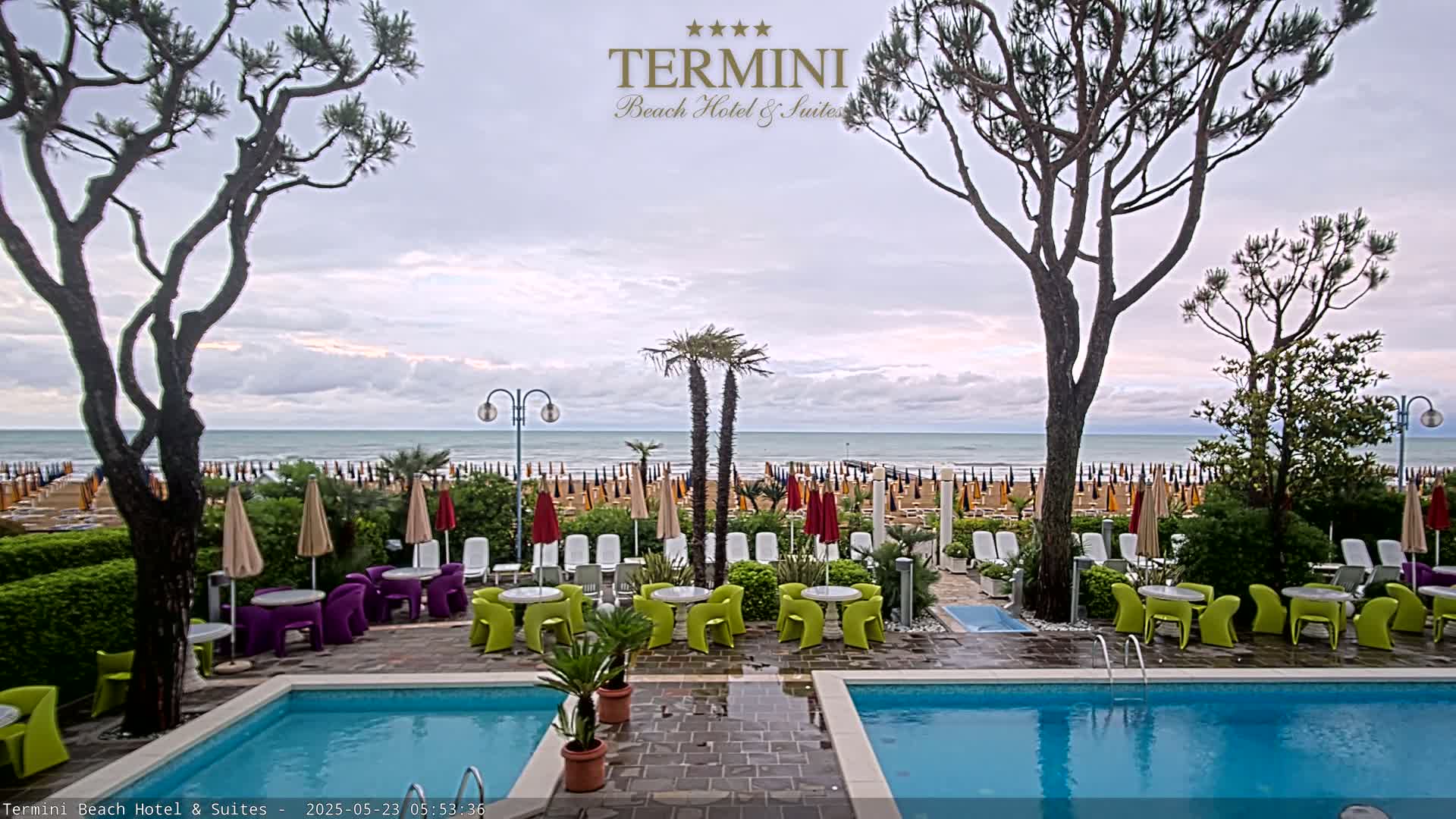A hotel pool area with lounge chairs and tables overlooks a beach with numerous umbrellas under an overcast sky.