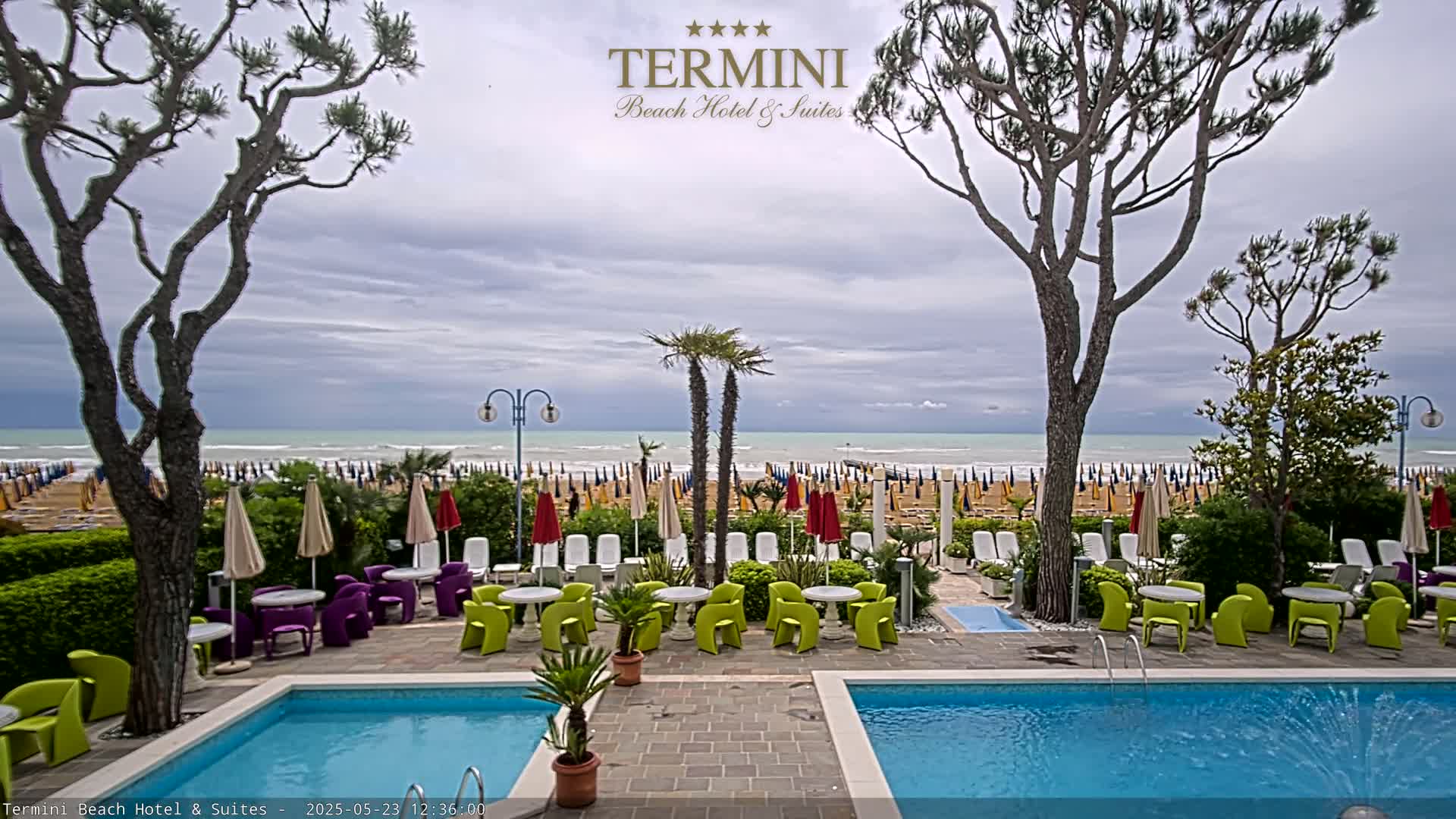 A hotel pool area with colorful patio furniture overlooks a beach with numerous umbrellas under a cloudy sky.