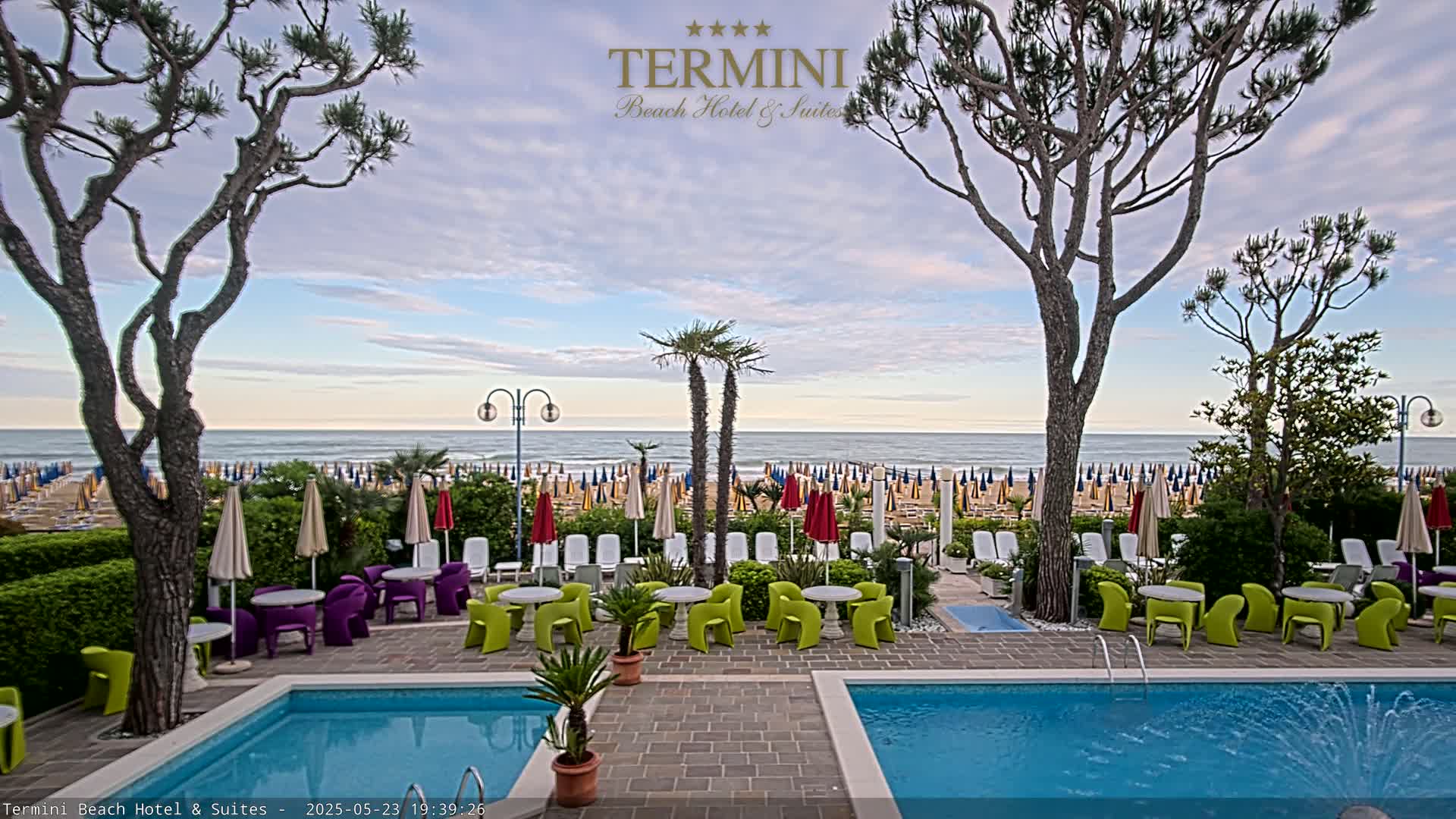 A hotel pool area with colorful patio furniture overlooks a beach dotted with umbrellas under a partly cloudy sky.