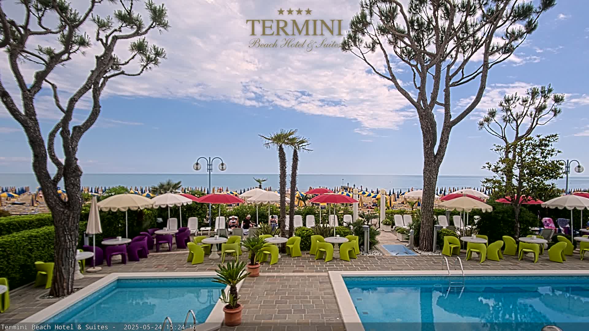 A hotel pool area with colorful patio furniture sits between two swimming pools, overlooking a beach with numerous umbrellas under a partly cloudy sky.
