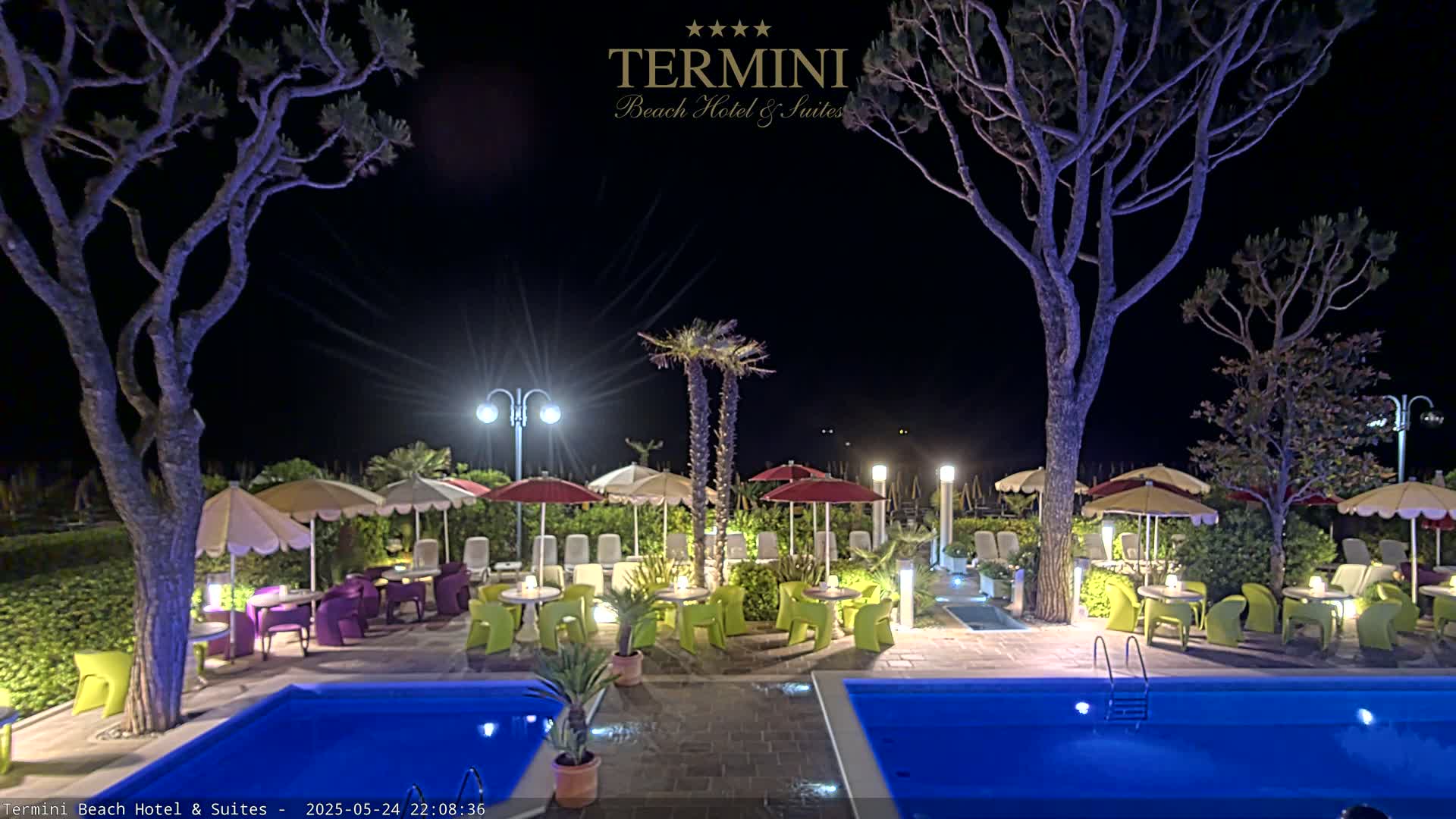 At night, an outdoor patio area with illuminated pools and colorful furniture under parasols is surrounded by trees and shrubs.