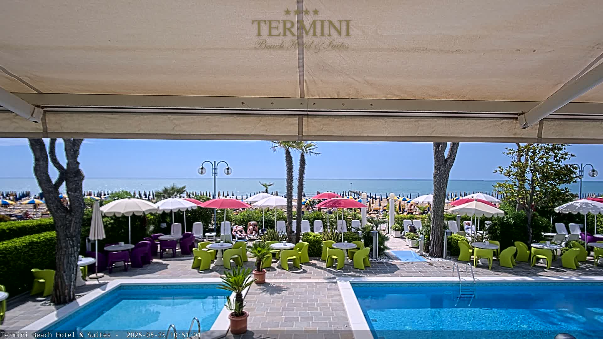 A sunny outdoor patio scene with two swimming pools, beach umbrellas, and seating areas overlooking a calm ocean on a bright, clear day.