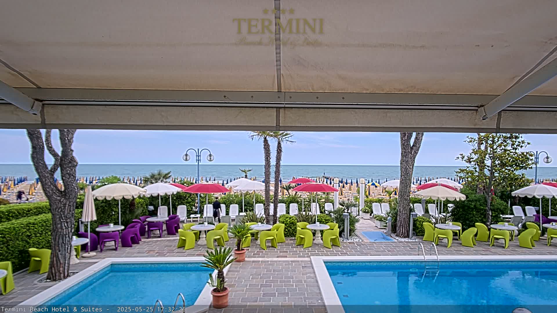 A view from under a patio awning shows a swimming pool, patio furniture, and beach umbrellas on a sandy beach with a calm ocean beyond.