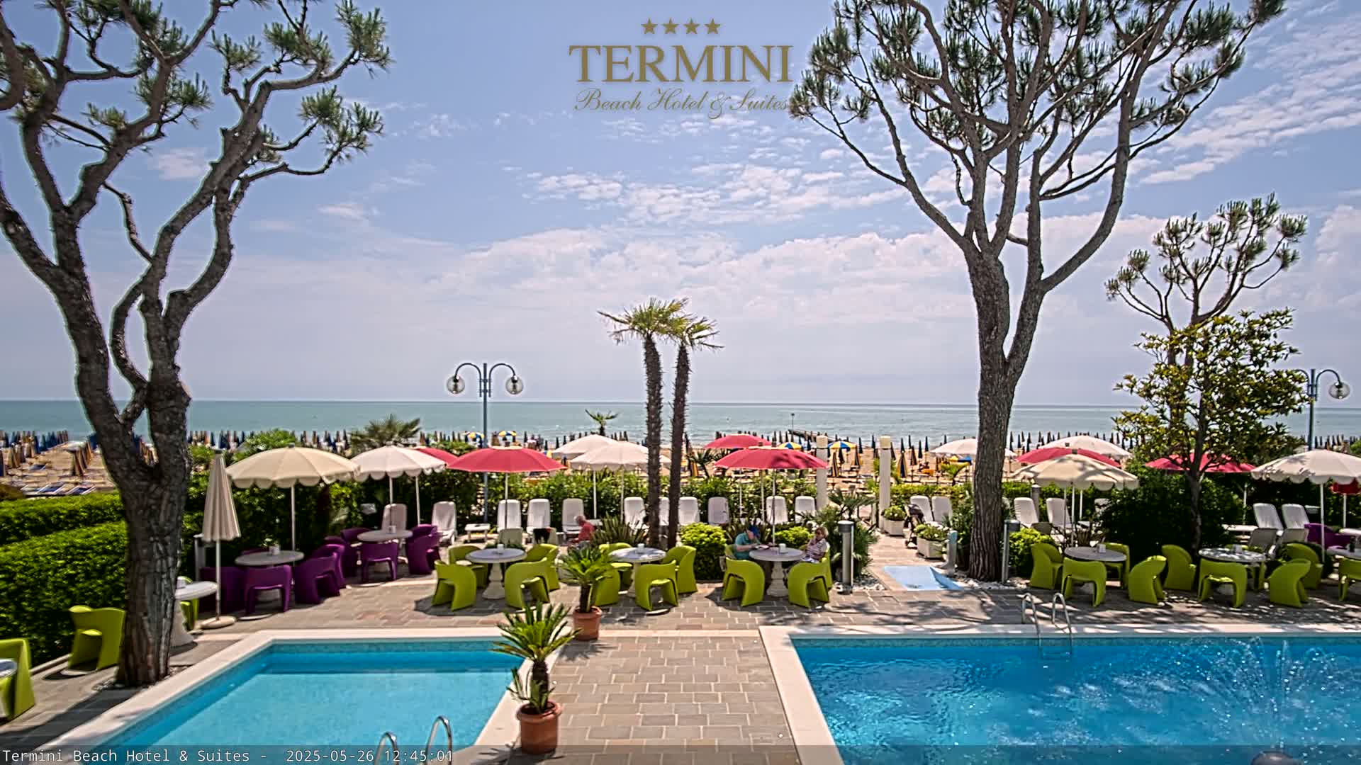 A resort pool area with colorful patio furniture and umbrellas overlooks a sunny beach and calm ocean.