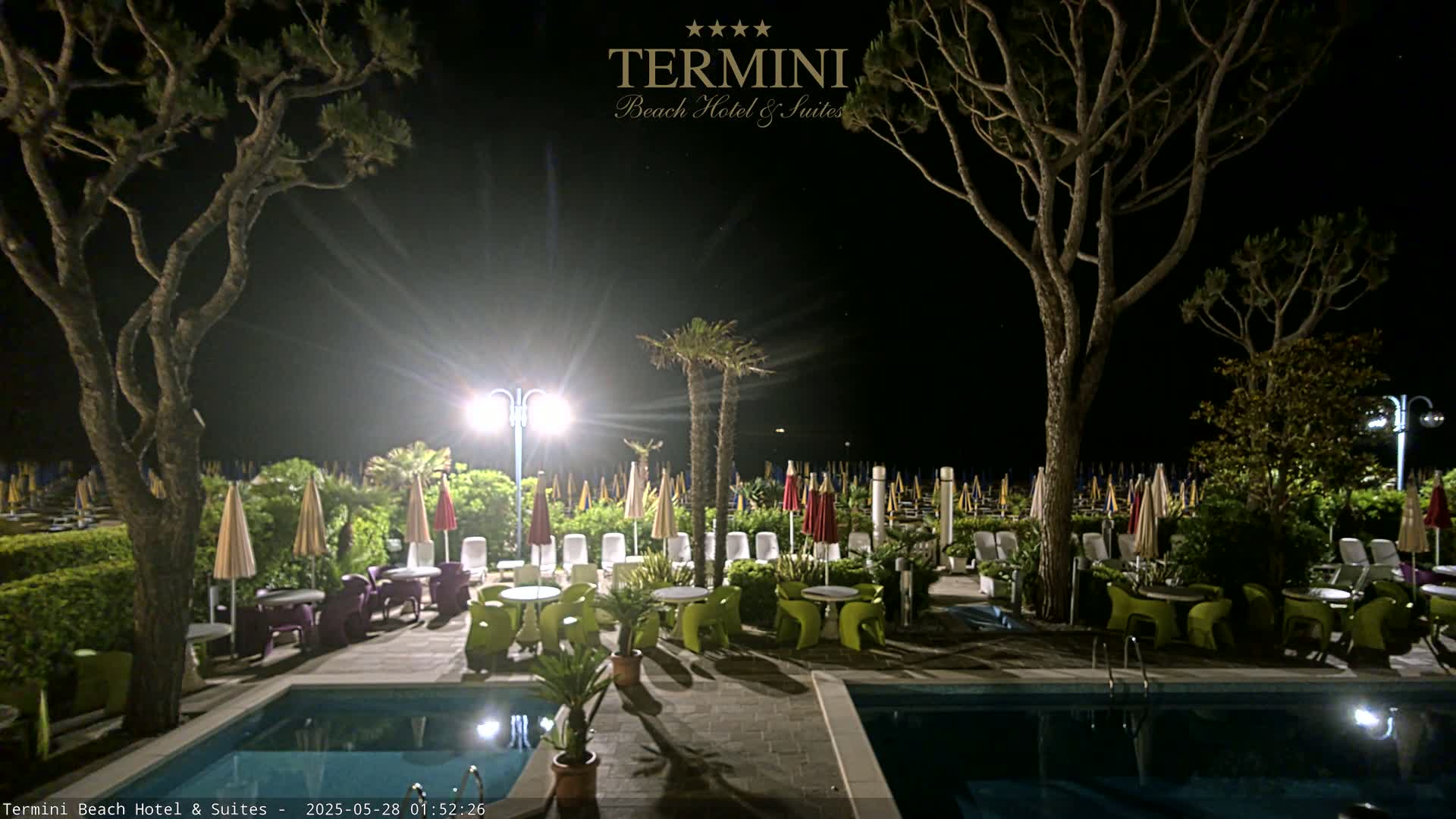 At night, a pool area with patio furniture under umbrellas is illuminated, surrounded by trees and adjacent to a beach with closed umbrellas.