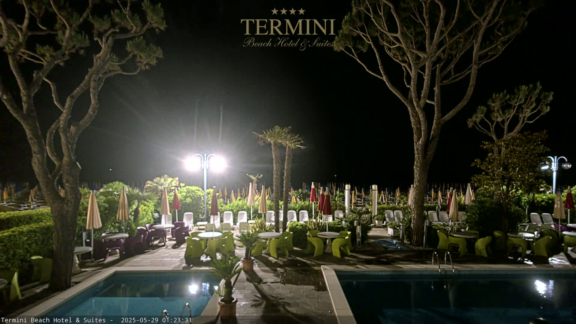 At night, an outdoor patio area with two swimming pools is illuminated, featuring lounge chairs, tables with colorful chairs, and tall trees under a dark sky.