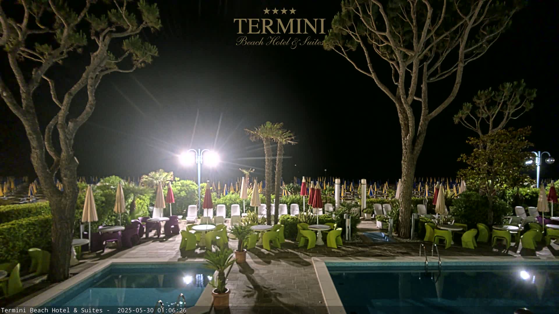 At night, a patio area with two pools and brightly lit tables and chairs sits between tall, dark trees, overlooking a beach with numerous closed umbrellas.