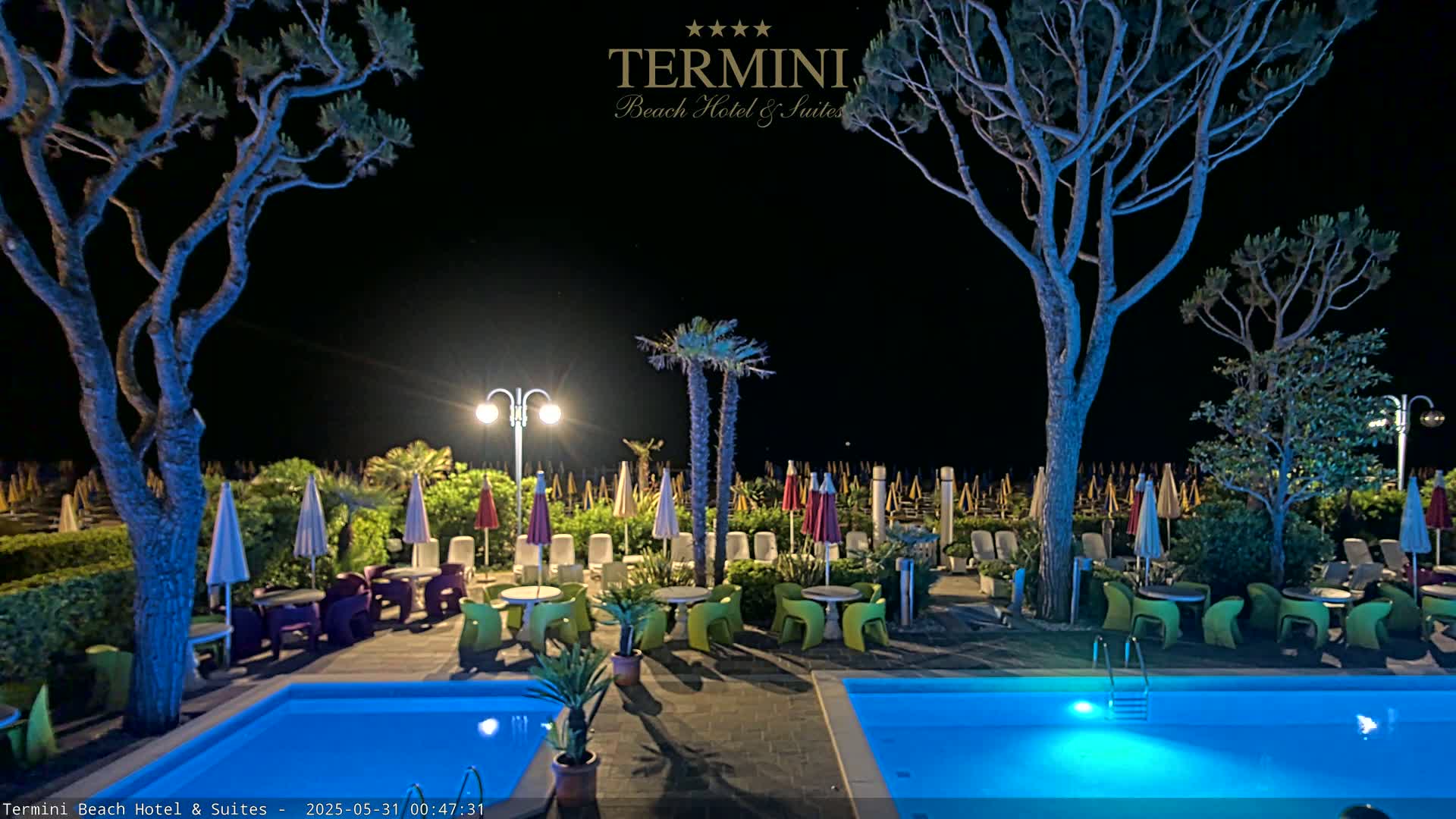 At night, a brightly lit resort area with two swimming pools, outdoor seating under umbrellas, and tall trees is visible.