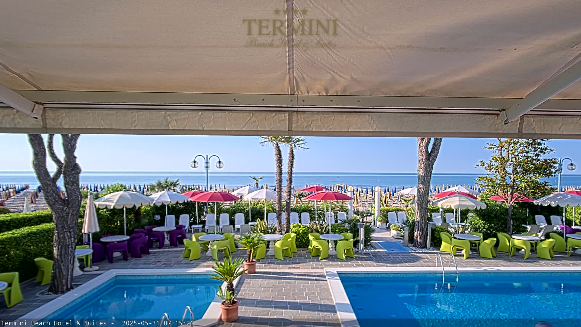 A sunny outdoor scene shows a swimming pool and patio area with colorful chairs and umbrellas overlooking a beach and calm ocean.