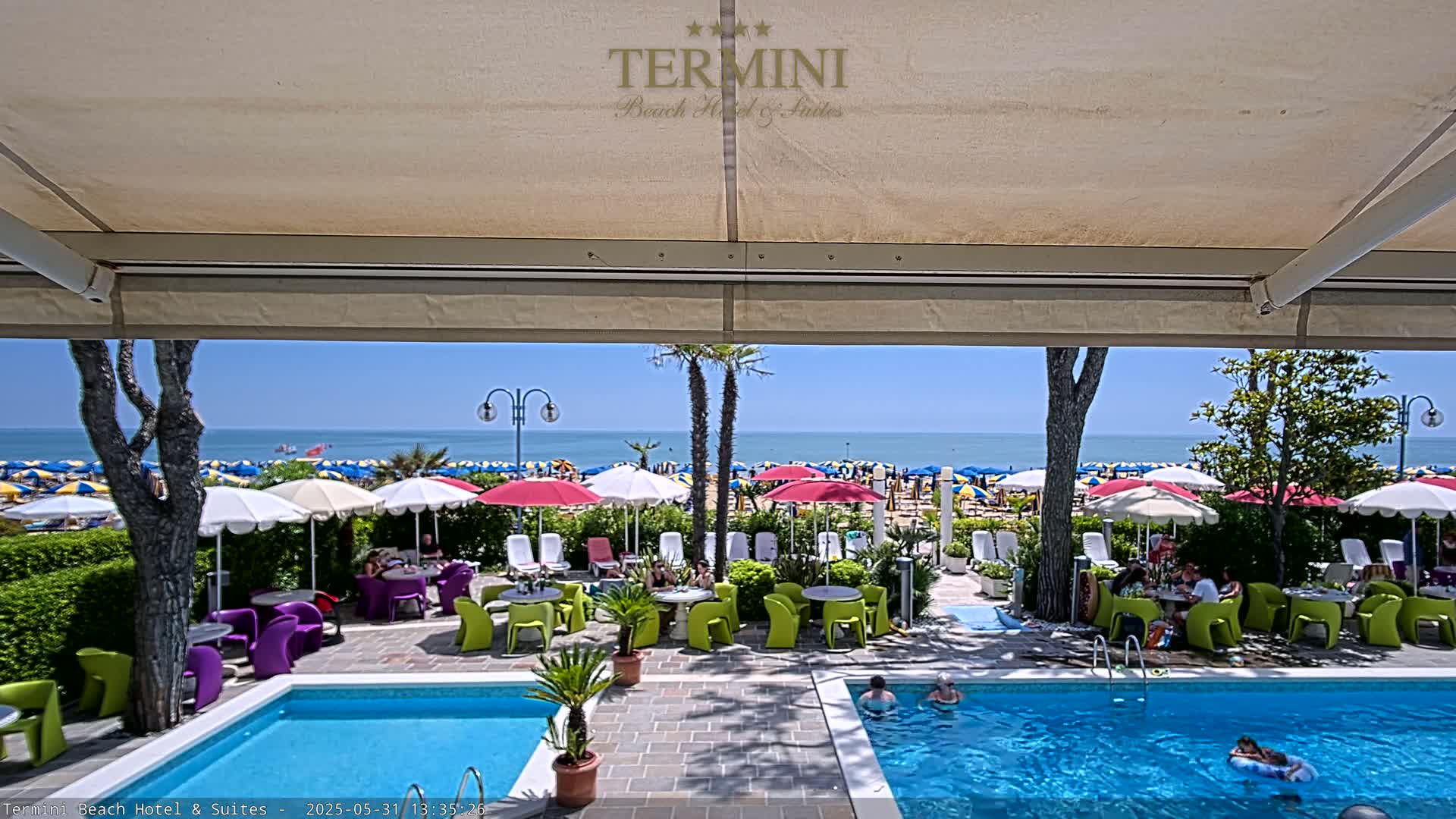 A patio with a swimming pool overlooks a sunny beach dotted with colorful umbrellas and a calm ocean.