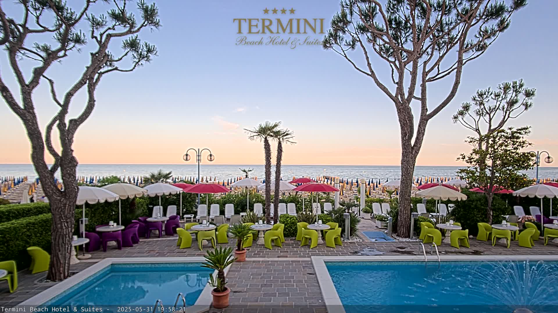 A hotel patio with a swimming pool overlooks a beach with numerous umbrellas under a sunset sky.