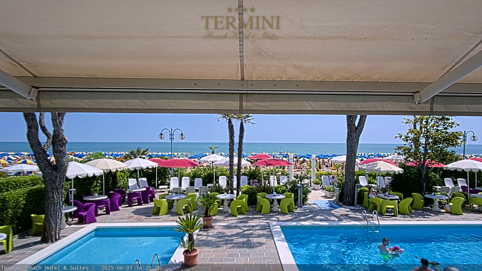 A view from under a patio awning shows a swimming pool, outdoor seating area, and a beach with numerous umbrellas under a clear blue sky.