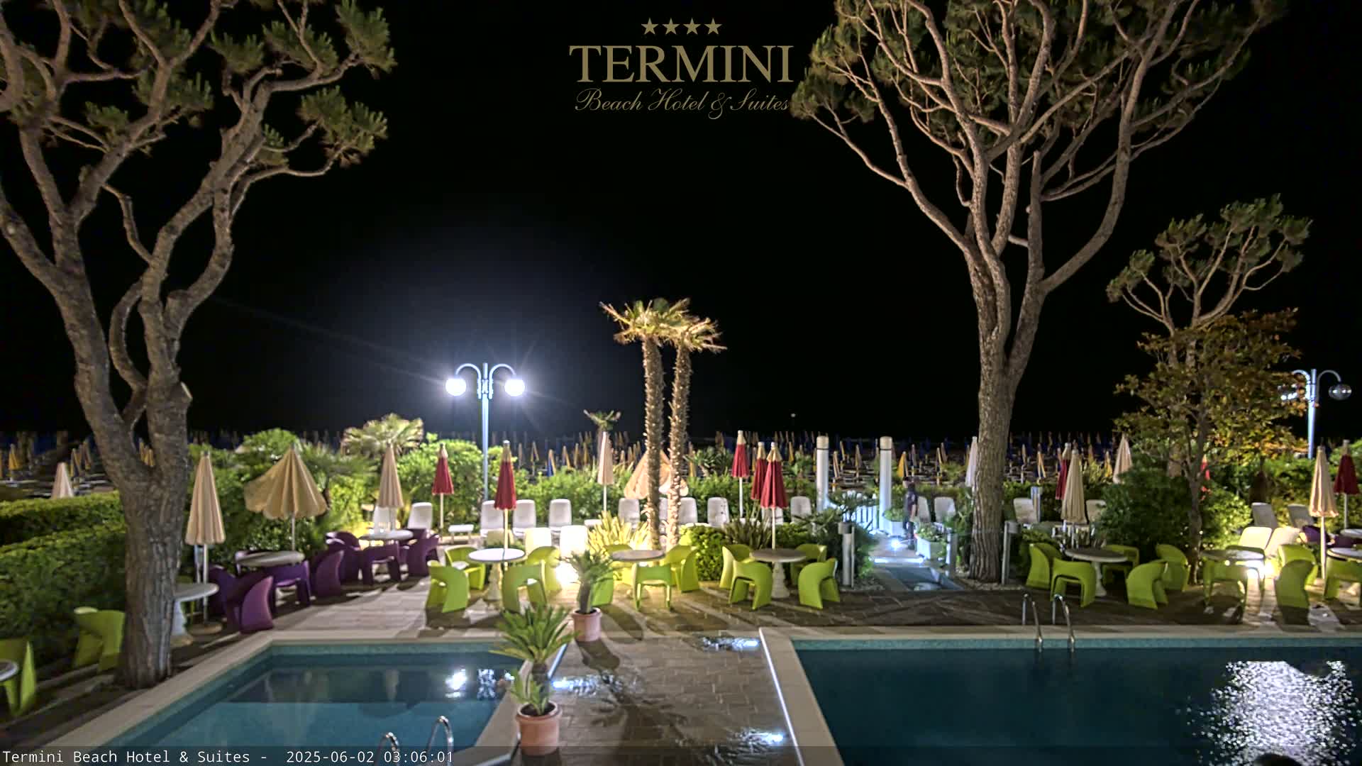 A nighttime view of a poolside patio area with tables, chairs, and umbrellas illuminated under outdoor lighting, surrounded by trees and palm trees.