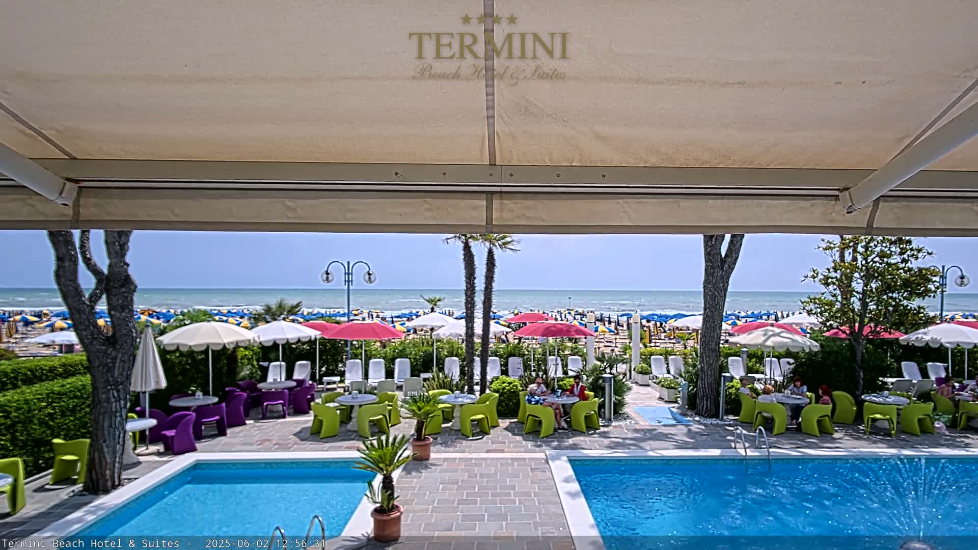 A sunny day at a beachside hotel features two pools, outdoor seating with colorful furniture under a retractable awning, and a view of a crowded beach with numerous umbrellas.