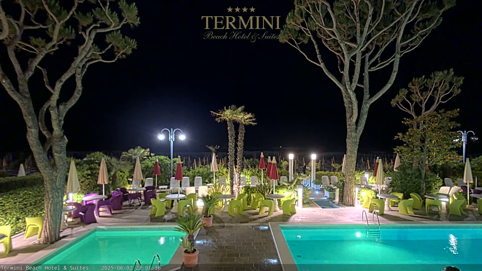At night, a pool area with illuminated tables and chairs under umbrellas is surrounded by trees and palm trees next to a brightly lit swimming pool.