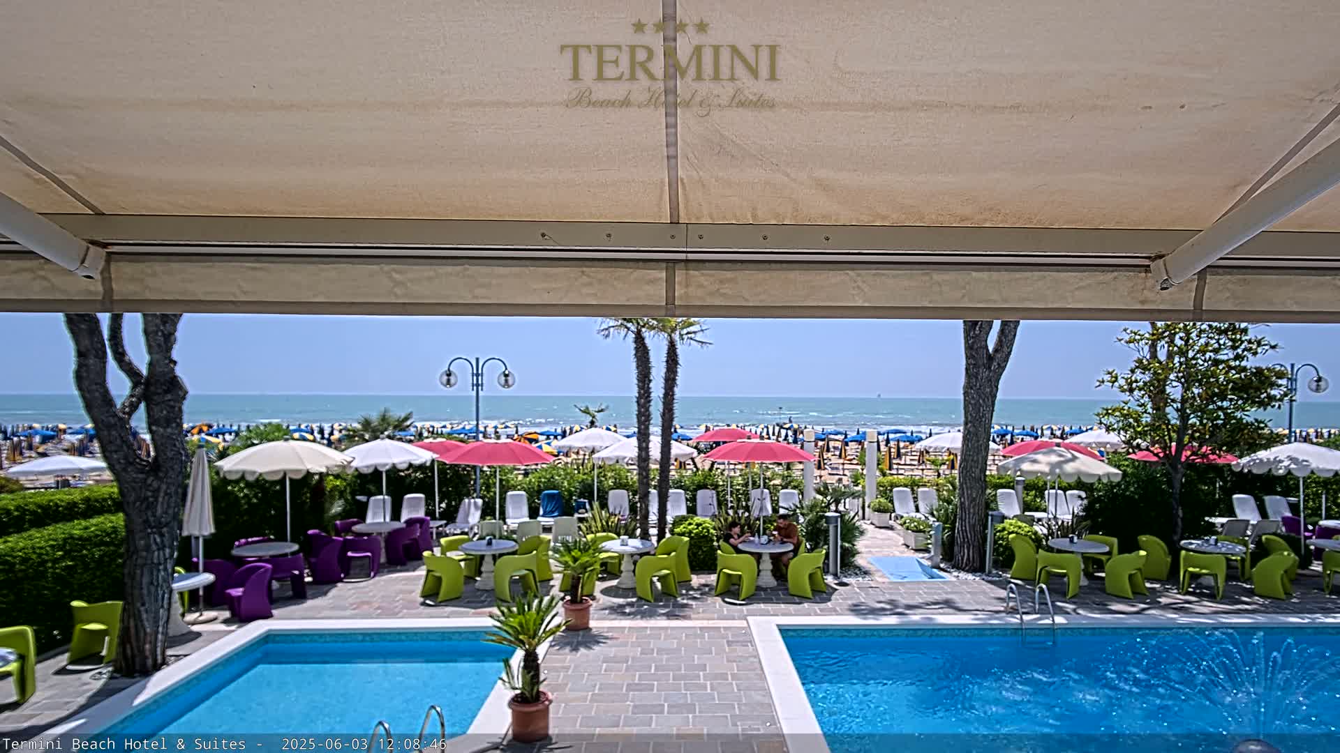 A view from under a patio awning shows a swimming pool, outdoor seating areas with colorful chairs and umbrellas, and a sunny beach with many people and umbrellas beyond.