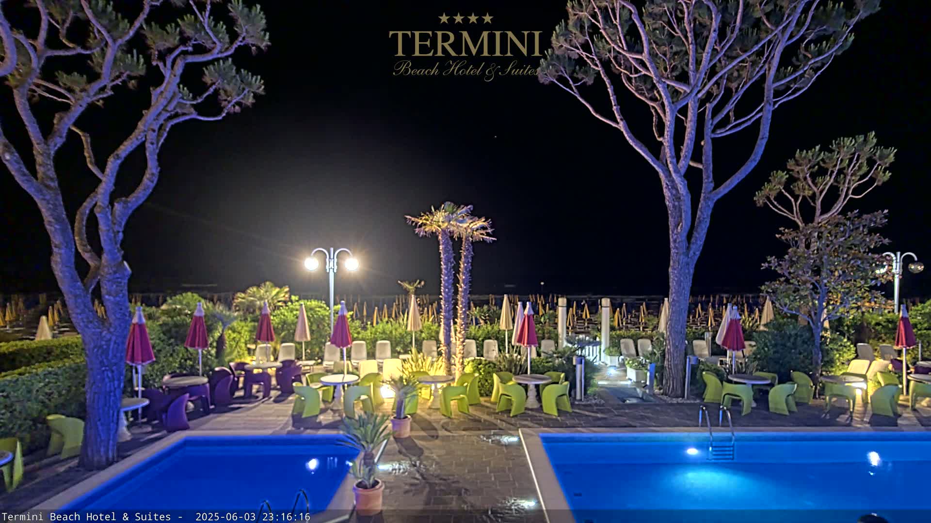 At night, a poolside patio with colorful furniture and umbrellas is illuminated under the dark sky near a beach with rows of closed umbrellas.