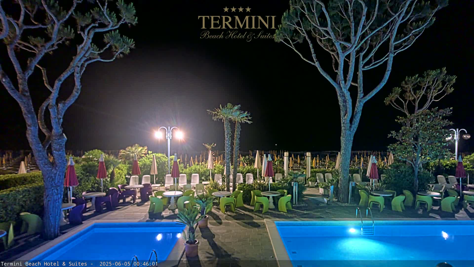 At night, a poolside patio area with two illuminated swimming pools, lounge chairs under umbrellas, and potted plants is surrounded by trees under a clear, dark sky.