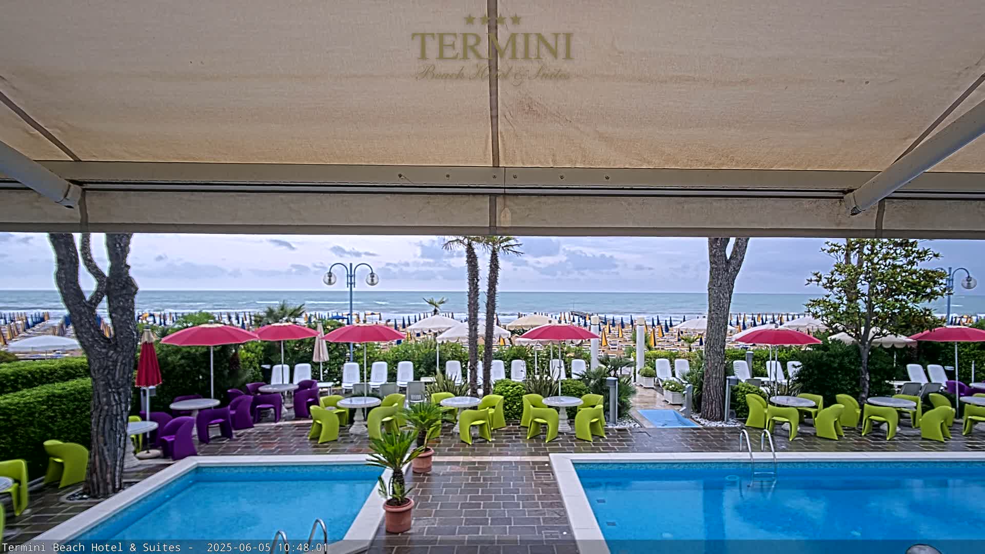 A view from under a patio awning shows two swimming pools, outdoor furniture, and a beach with numerous umbrellas under an overcast sky.
