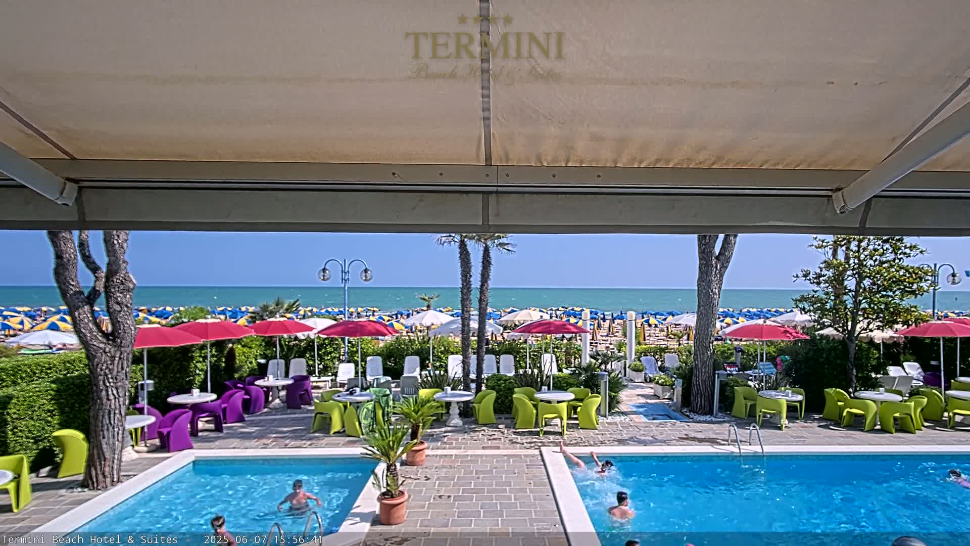 A view from under a patio awning shows a swimming pool with people swimming, patio furniture, and beyond that, a beach with numerous colorful umbrellas under a bright, sunny sky.