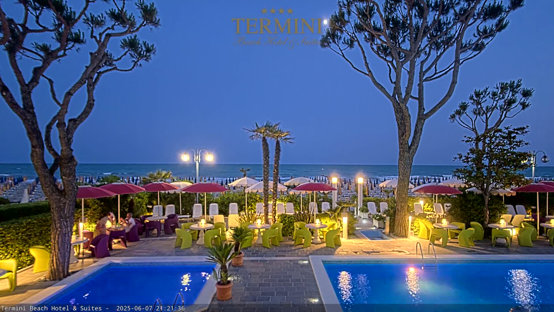 A twilight scene shows two swimming pools and an outdoor seating area with umbrellas and tables near a calm ocean under a clear, dark-blue sky.