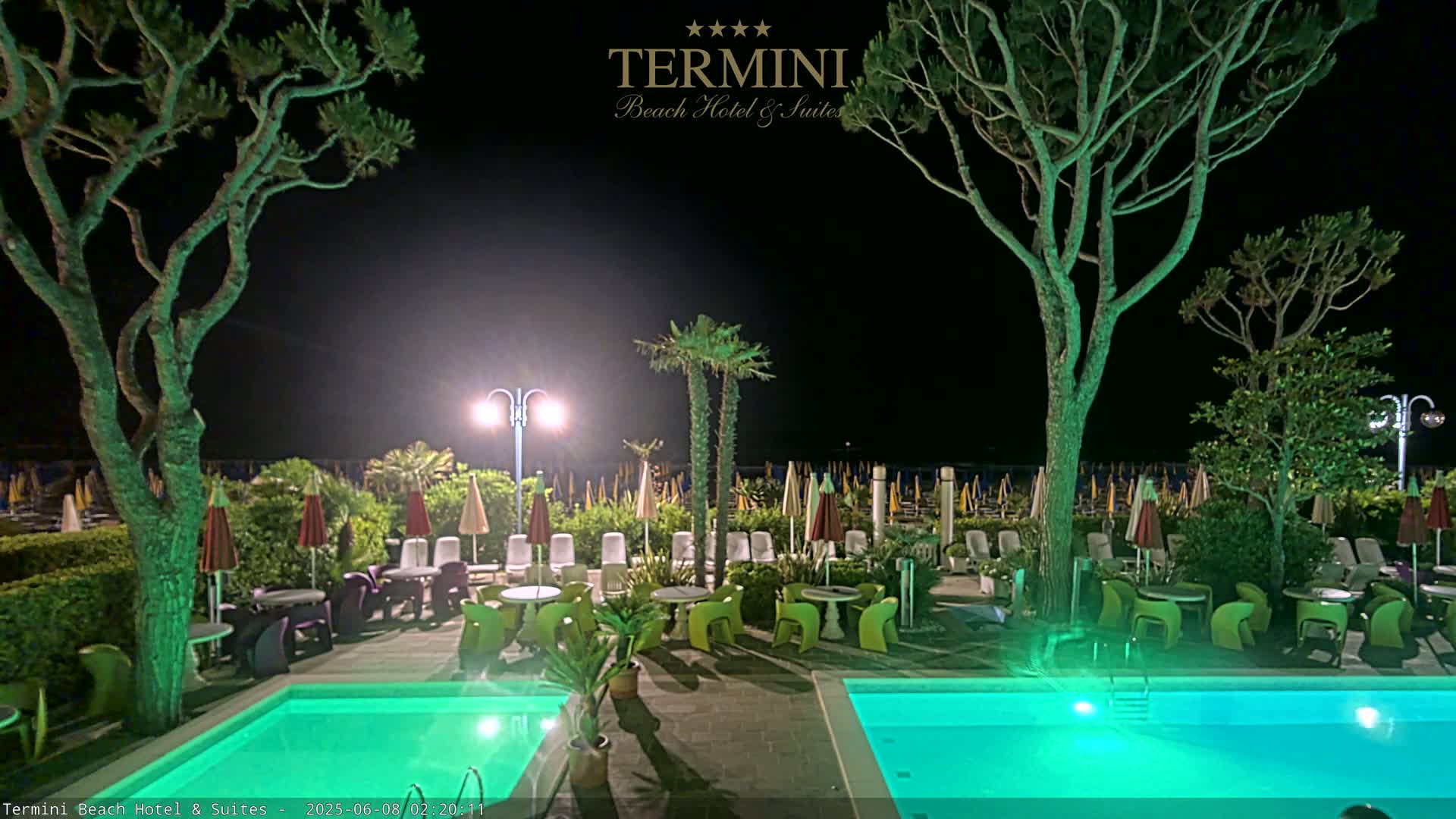 At night, a hotel pool area with illuminated green water, surrounded by trees, lounge chairs, and tables, overlooks a beach with numerous closed umbrellas.