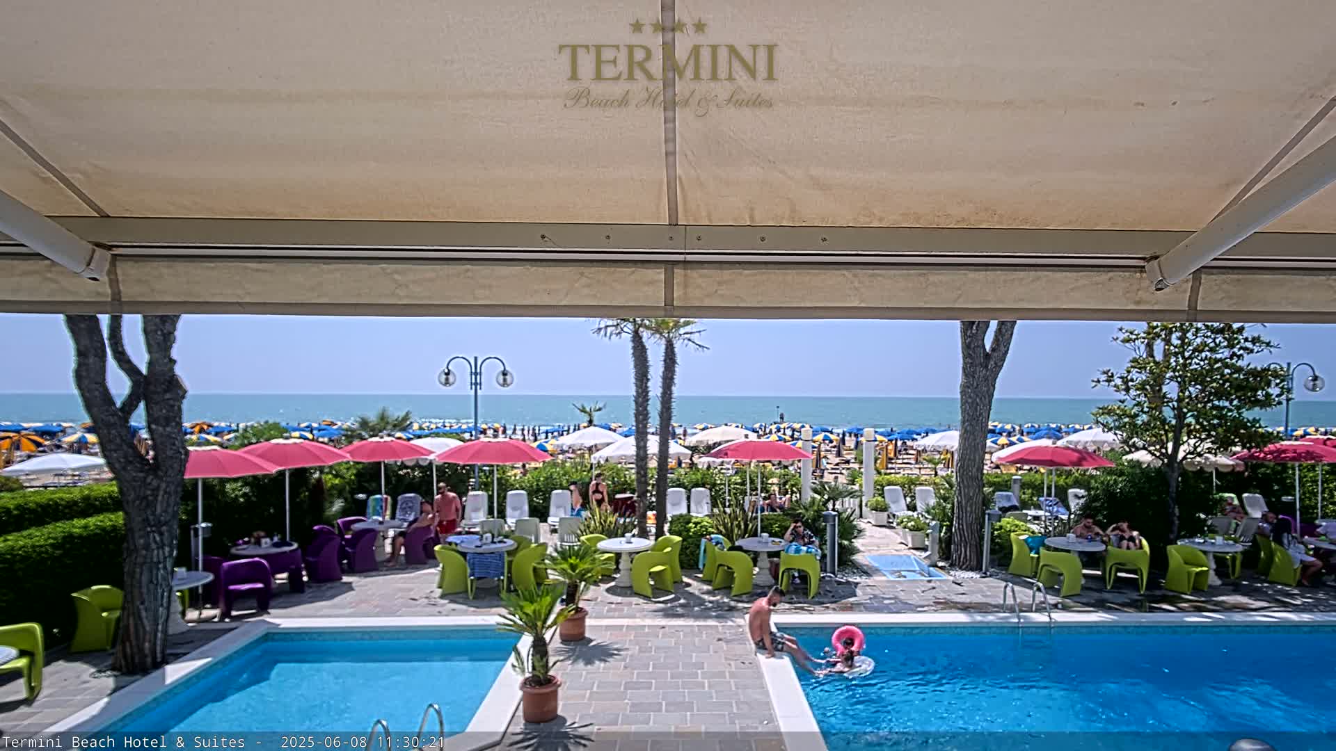 A sunny day at a beachside hotel with a pool, showing many beach umbrellas and people on a sandy beach beyond.