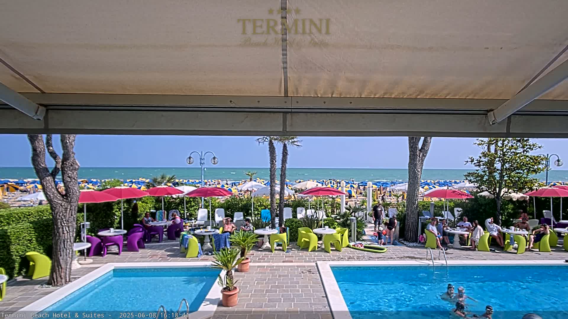 A sunny day at a beachfront hotel shows two swimming pools surrounded by people sitting at tables under umbrellas, with a view of a beach crowded with more umbrellas and people.