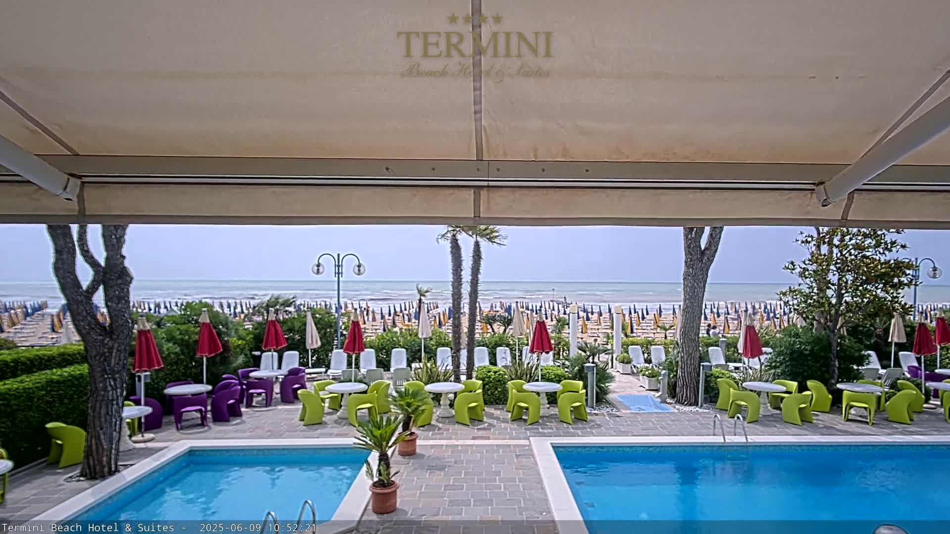 A partially covered patio area with a pool overlooks a beach with numerous umbrellas and a calm ocean under a partly cloudy sky.