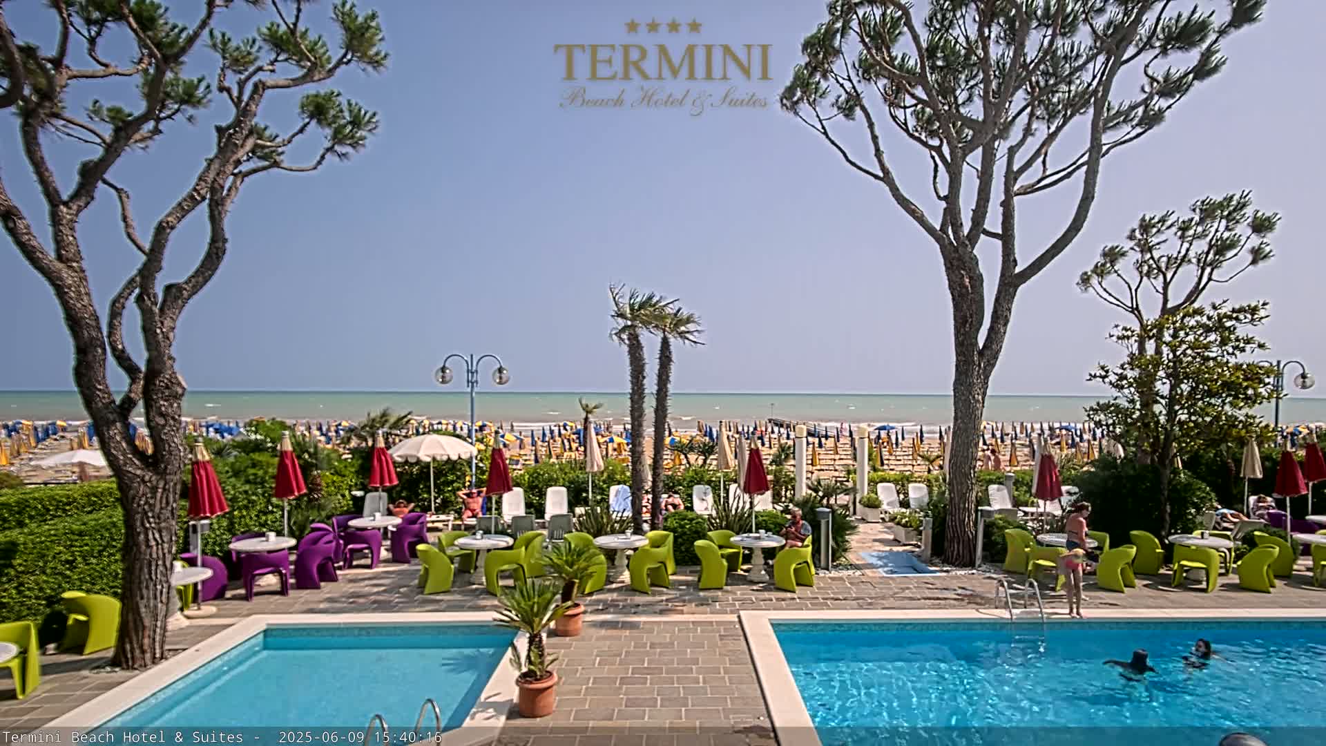 A resort pool area with bright green and purple furniture sits between two swimming pools, overlooking a sunny beach with numerous umbrellas under a clear blue sky.