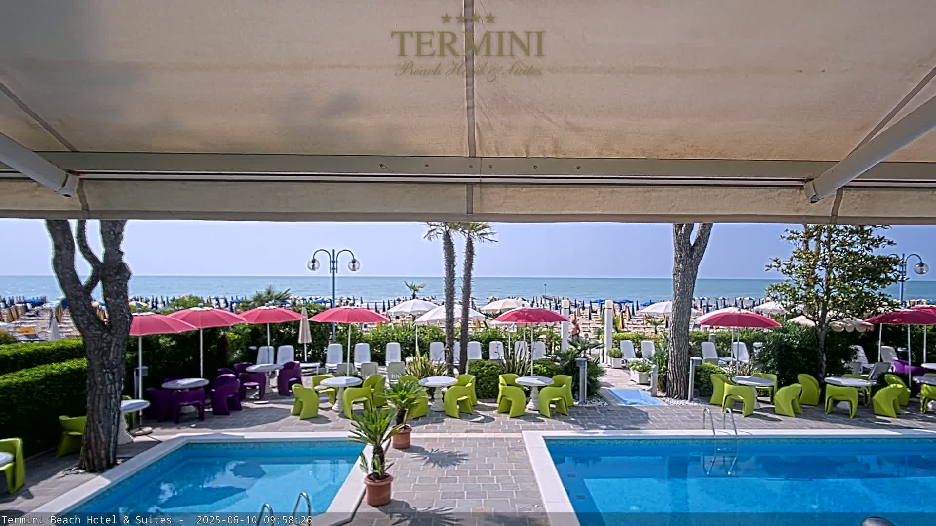 A view from under a patio awning shows a swimming pool, patio furniture, and a beach with numerous umbrellas and lounge chairs under a clear sky.