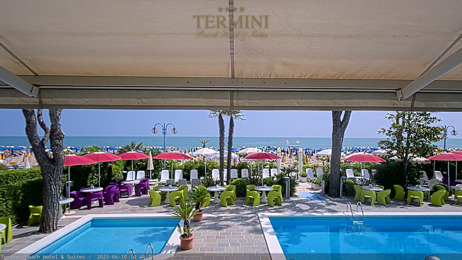 A view from under a patio awning shows two swimming pools and a patio with colorful chairs and umbrellas overlooking a sunny beach and ocean.