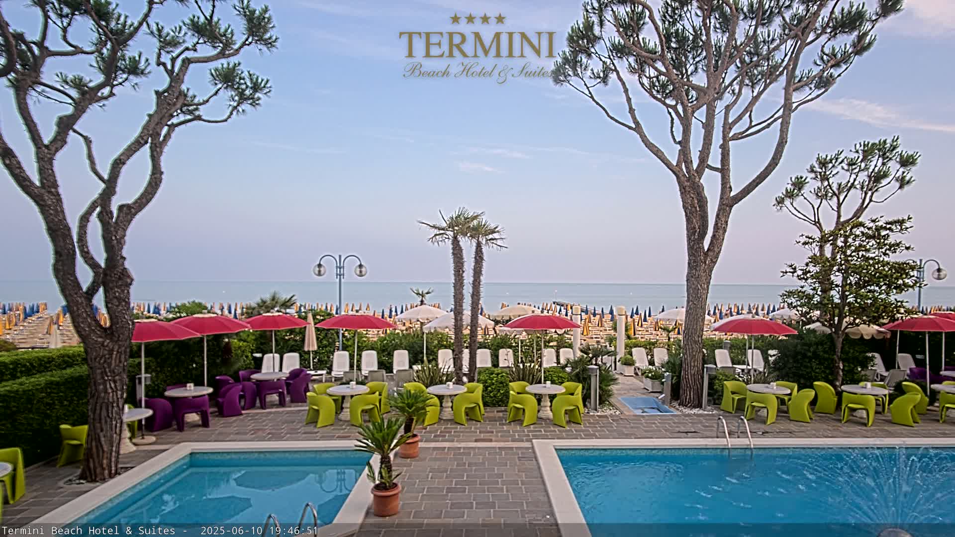 A resort pool area with umbrellas, tables, and chairs sits between two tall trees, overlooking a beach and calm ocean under a clear sky.