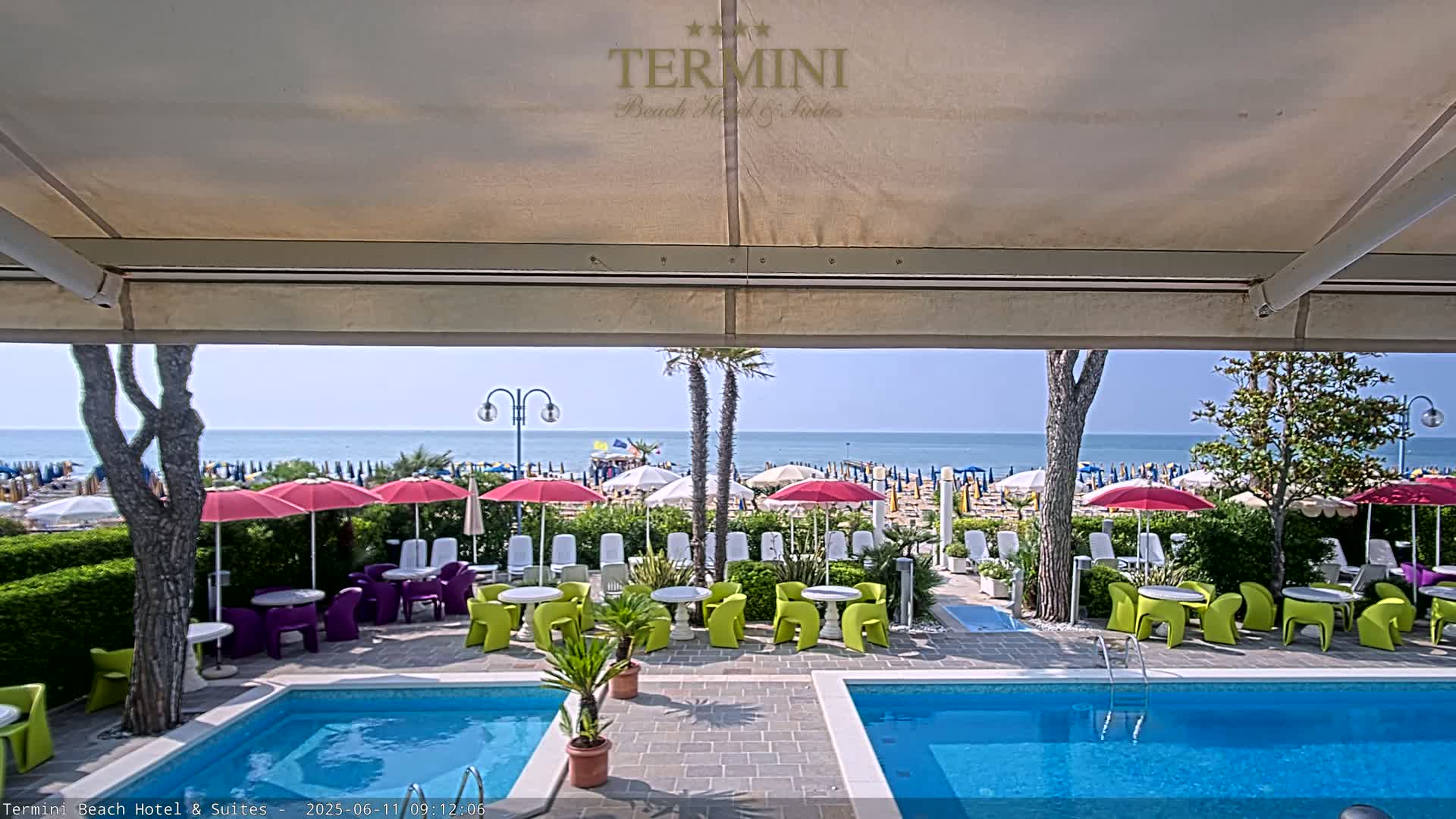 A patio with two swimming pools overlooks a sunny beach with numerous umbrellas and lounge chairs.