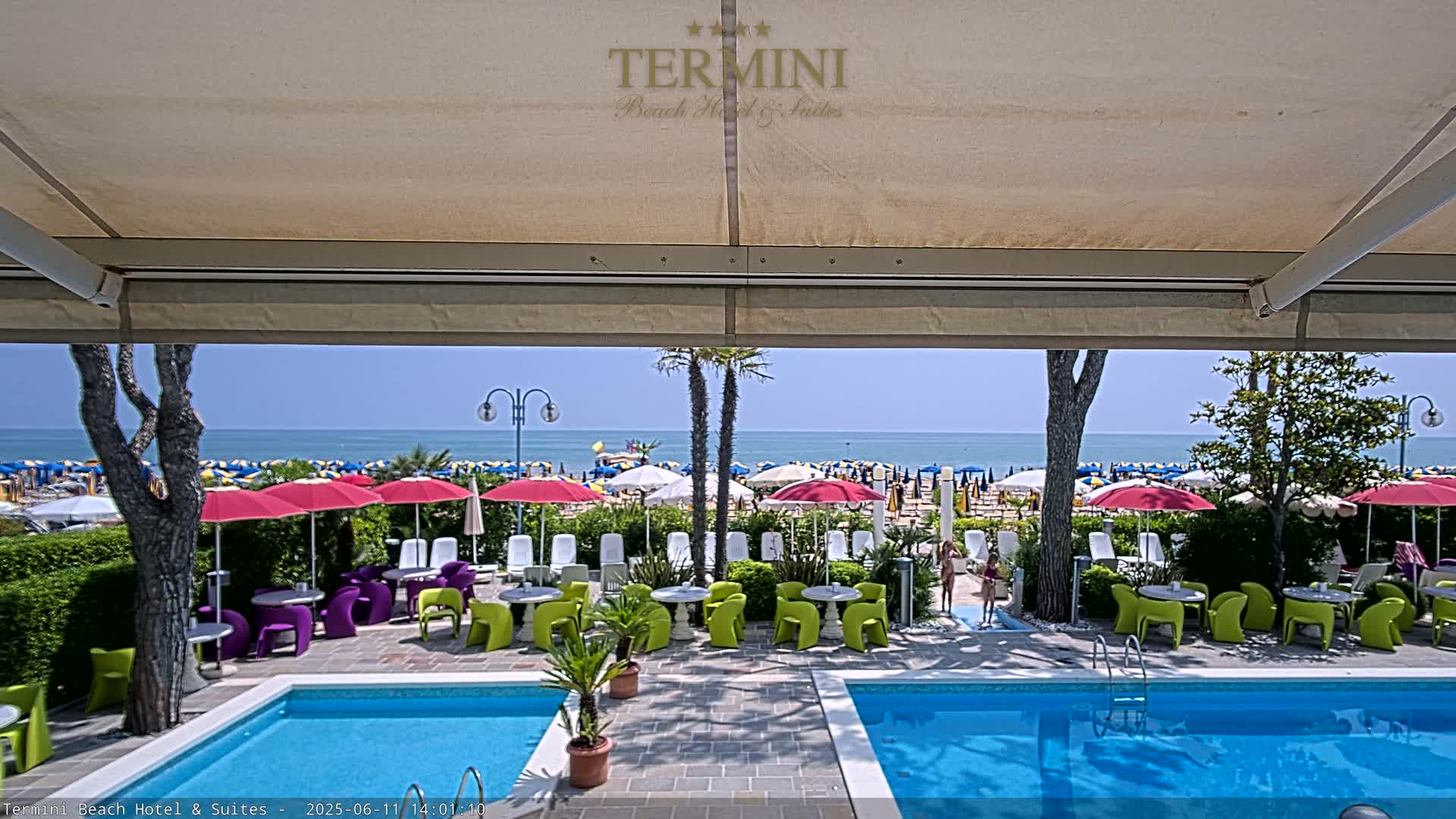 From under a patio awning, two swimming pools are visible, overlooking a sunny beach crowded with umbrellas and people.