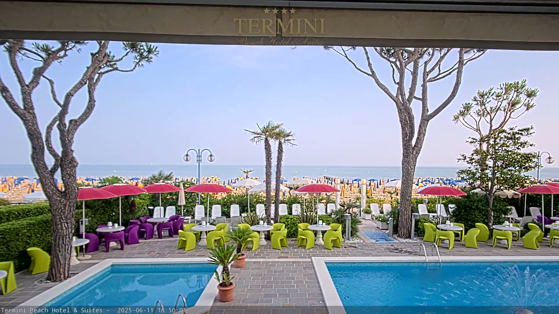 A patio with two swimming pools, colorful outdoor furniture, and umbrellas overlooks a beach with numerous beach umbrellas under a clear sky.