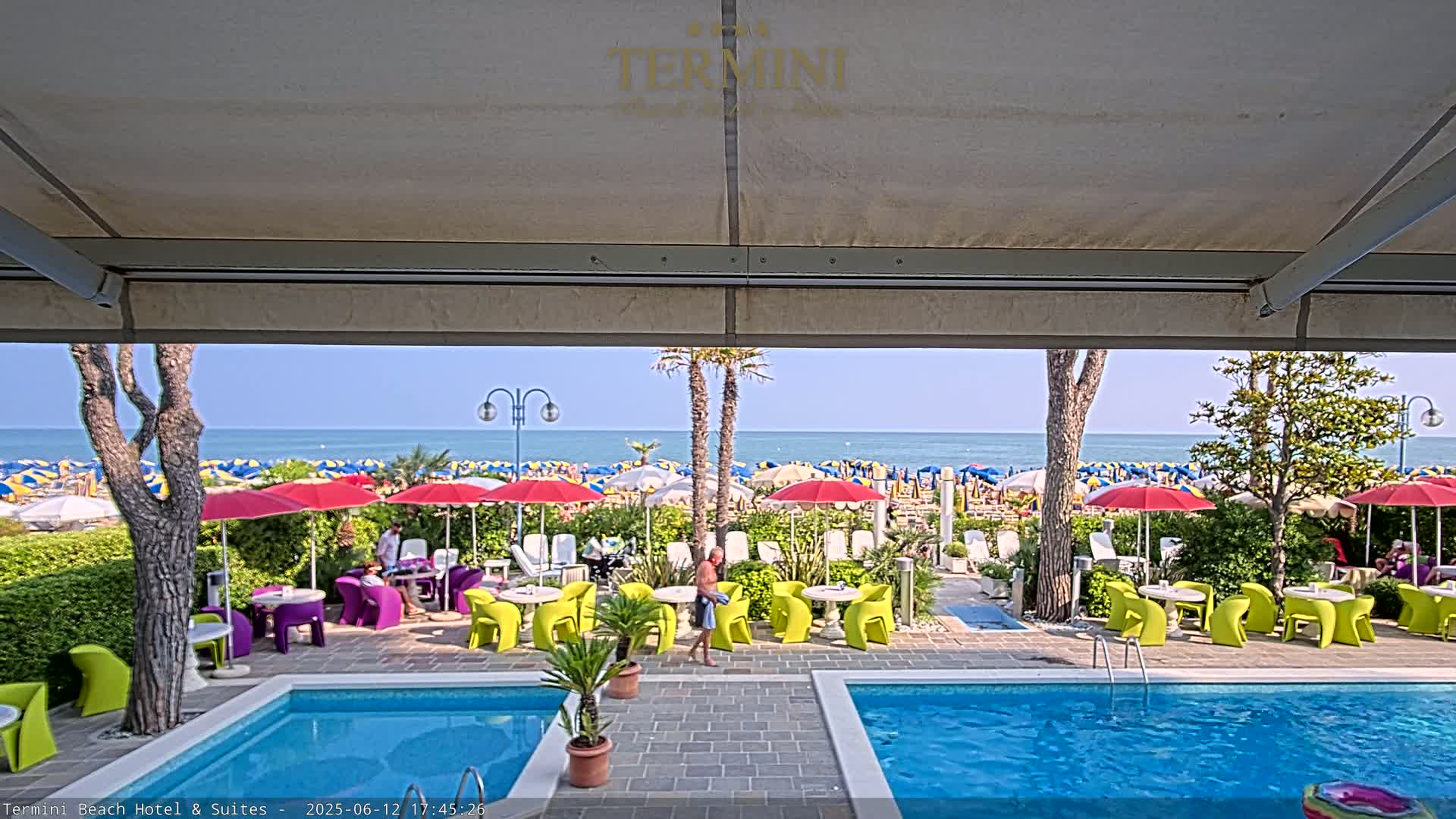A view from under a patio awning shows a sunny beach with colorful umbrellas and lounge chairs, beyond a patio with bright-colored chairs and tables surrounding two swimming pools.