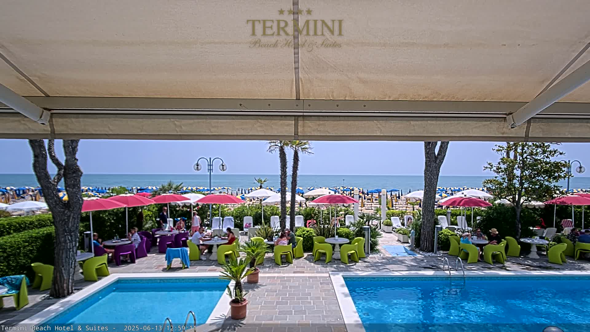 A sunny outdoor scene viewed from under a patio awning shows two swimming pools, a patio with tables and chairs under umbrellas, and a beach with many umbrellas in the background.
