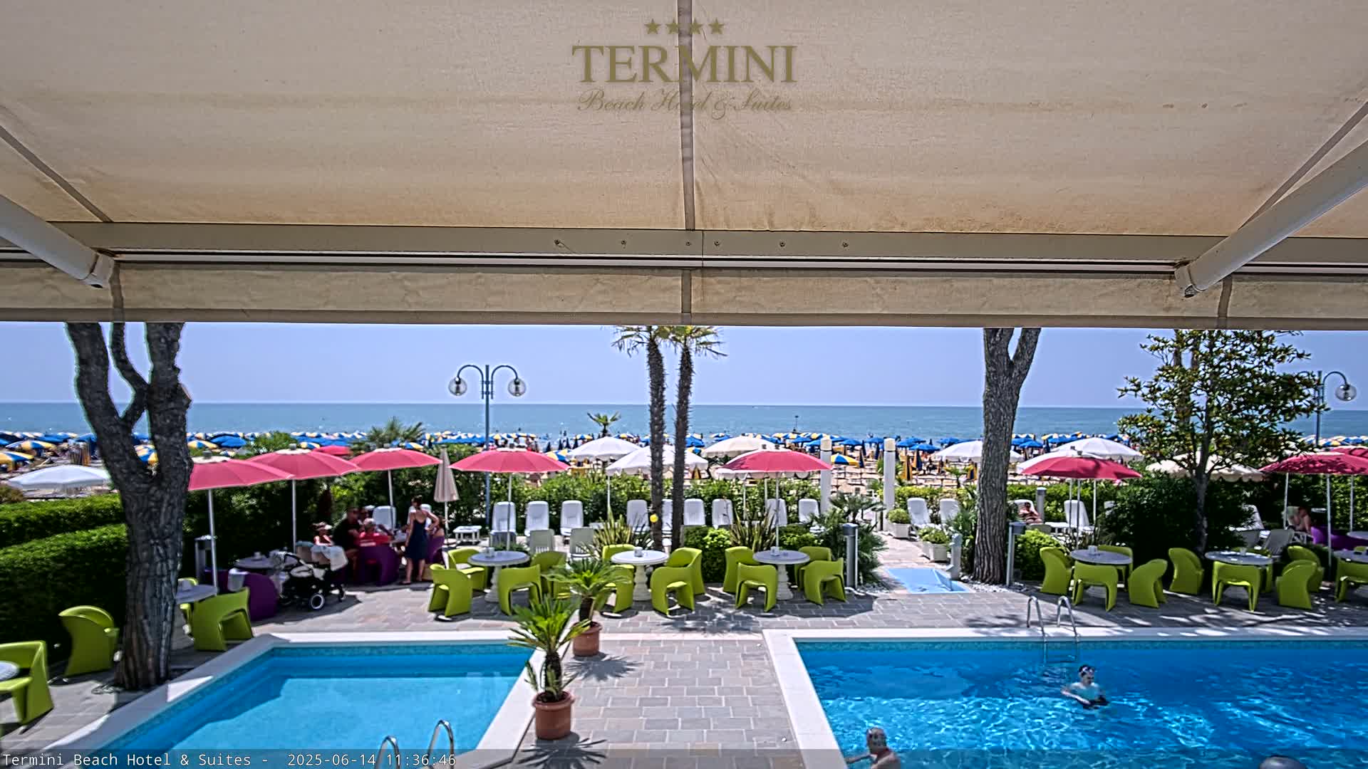 A sunny outdoor scene shows two swimming pools and a patio area with brightly colored tables and chairs, all overlooking a beach filled with umbrellas and people.