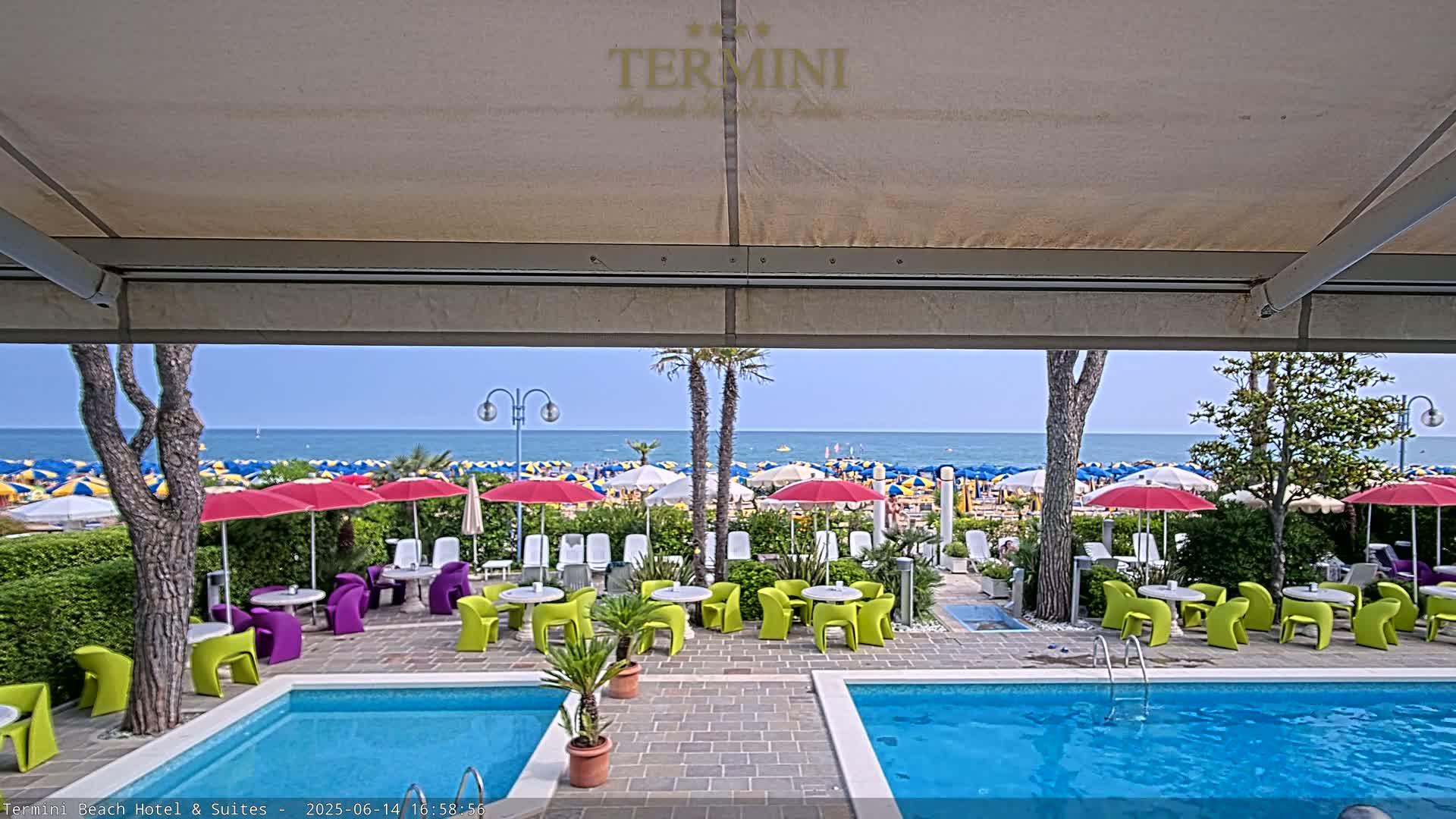 A view from under a patio awning shows a patio with two swimming pools, brightly colored outdoor furniture, and many beach umbrellas on a sandy beach beyond, all under a clear blue sky.