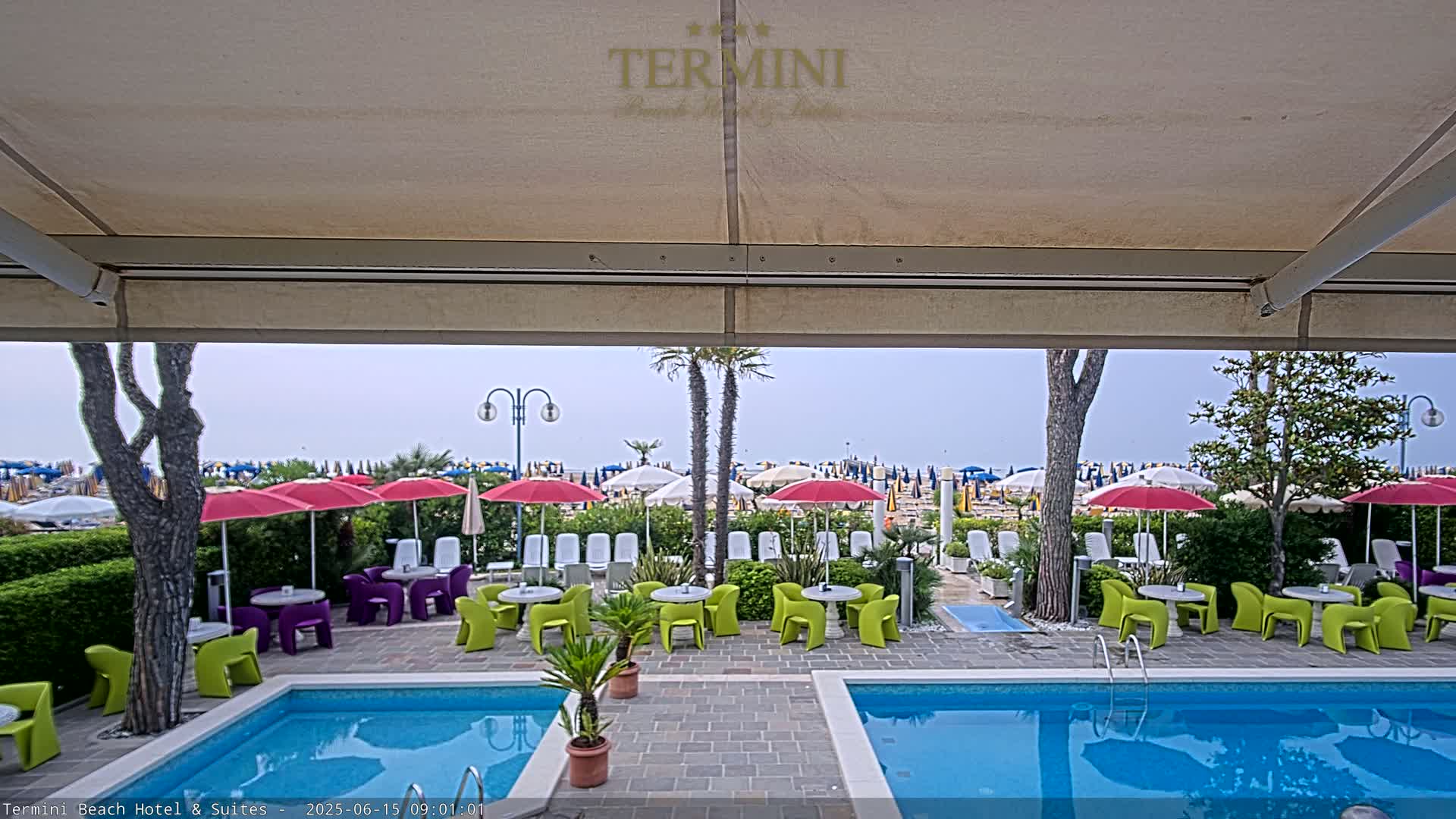 A view from under a patio awning shows a swimming pool and patio furniture, beyond which are beach umbrellas and lounge chairs on a hazy day.