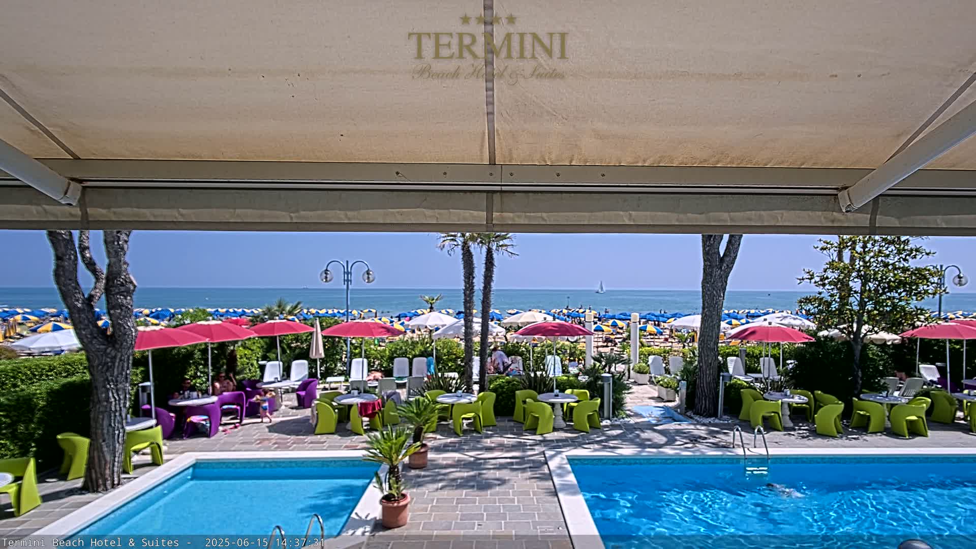 A sunny outdoor patio scene with two swimming pools, colorful patio furniture, and beach umbrellas on a sandy beach visible in the background.
