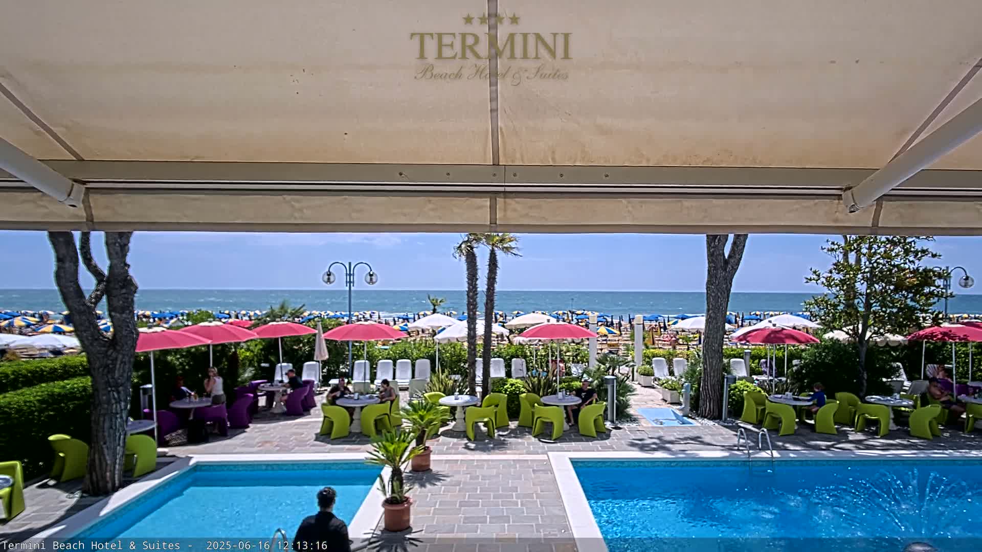 A sunny day at a resort shows two swimming pools, outdoor seating areas with people relaxing, and a beach with numerous umbrellas under a clear sky.