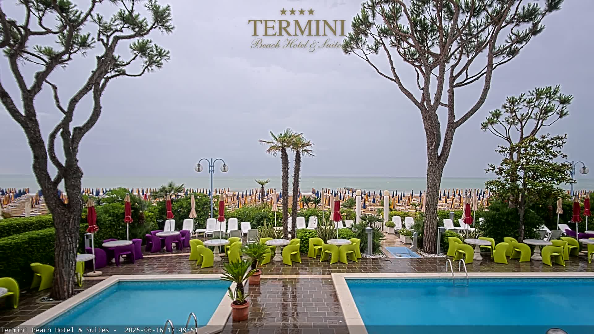 A patio with two swimming pools overlooks a beach with numerous umbrellas under an overcast sky.