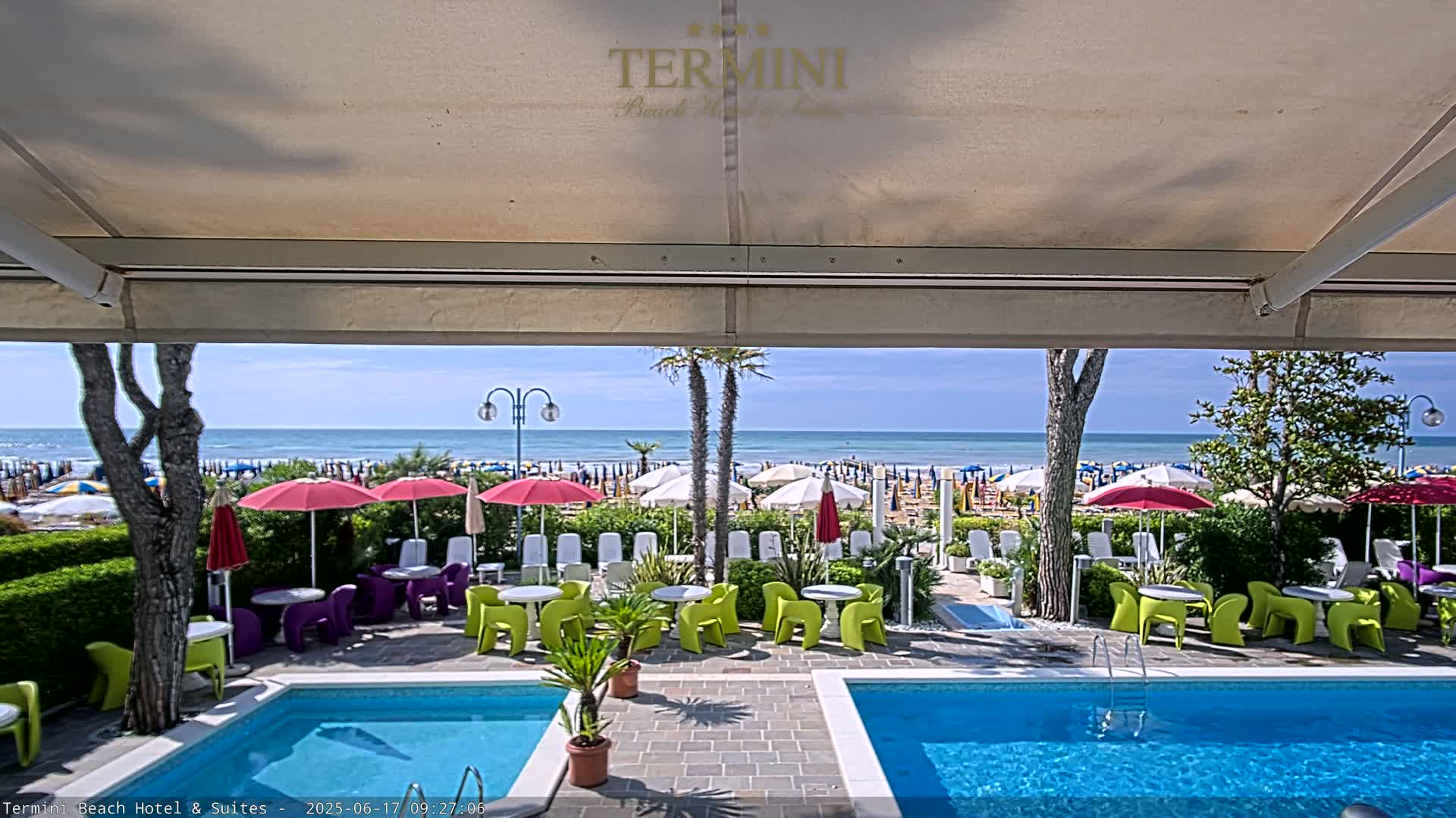 Two swimming pools sit beside a patio with brightly colored chairs and umbrellas, overlooking a sunny beach with calm ocean waves.