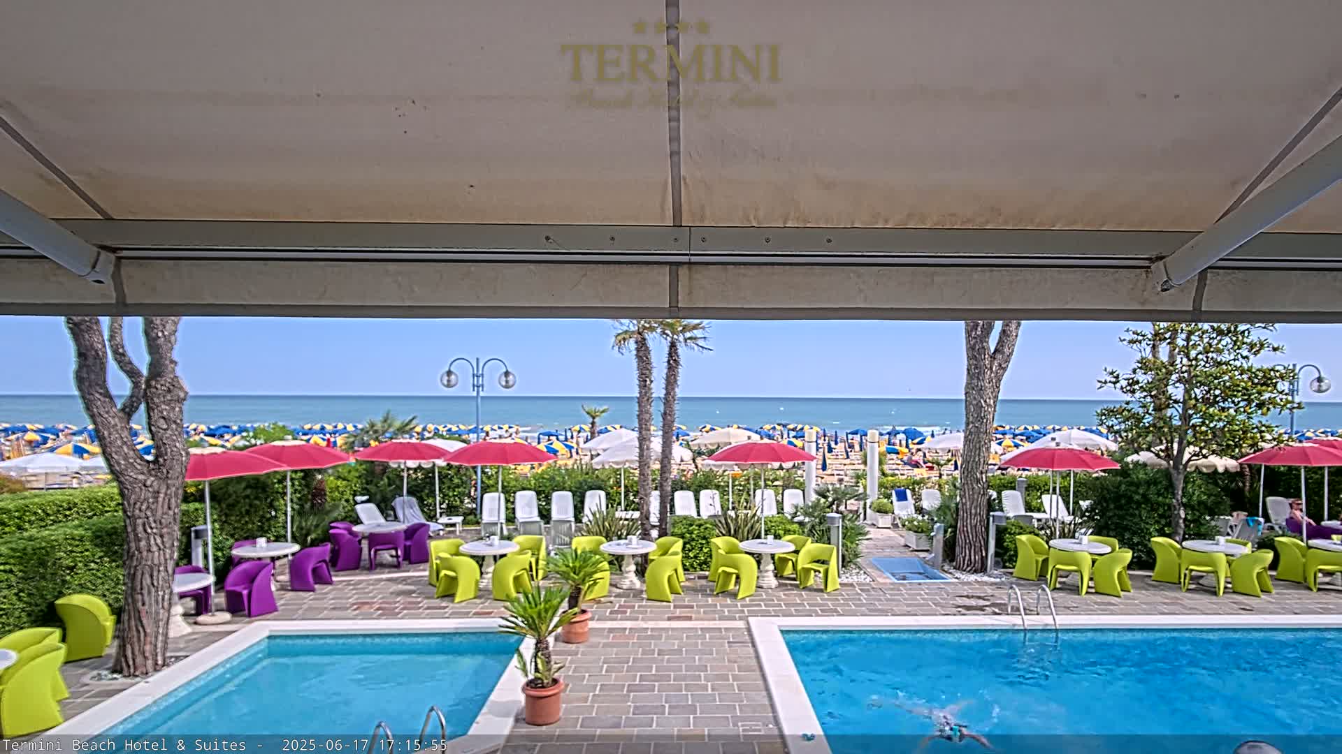 A view from under a patio awning shows a sunny beach with colorful umbrellas and lounge chairs, beyond a swimming pool area with bright green and purple outdoor furniture.
