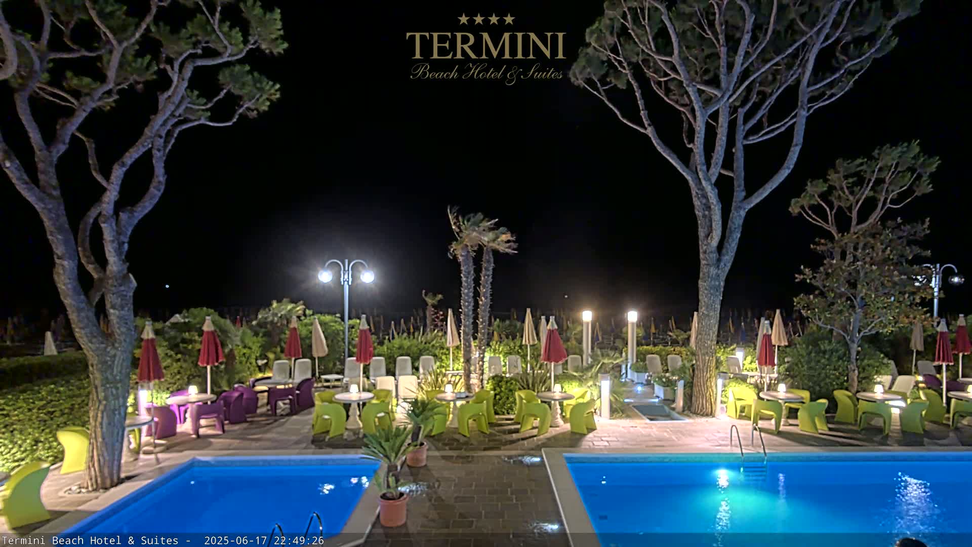 At night, a resort pool area with two illuminated pools, patio furniture under umbrellas, and palm trees is shown.